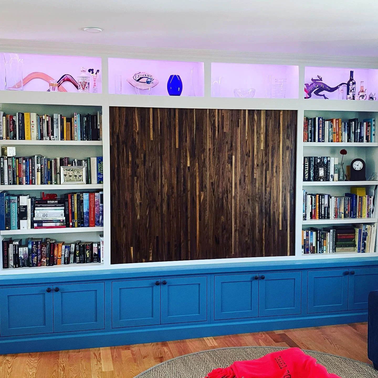 A built-in bookshelf with white framing, filled with books, decorative items, and a clock. The lower part of the bookshelf is painted blue with cabinet doors. The wall above the bookshelf has display shelves with glassware and decorative items, illum