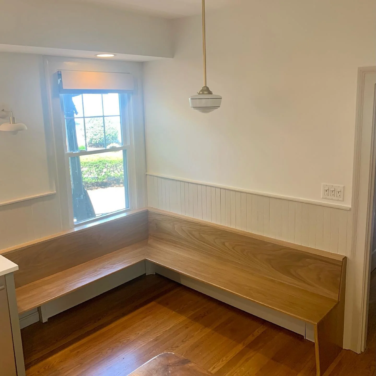 Corner of a kitchen with a built-in wooden bench, a window showing greenery outside, and a ceiling light fixture.