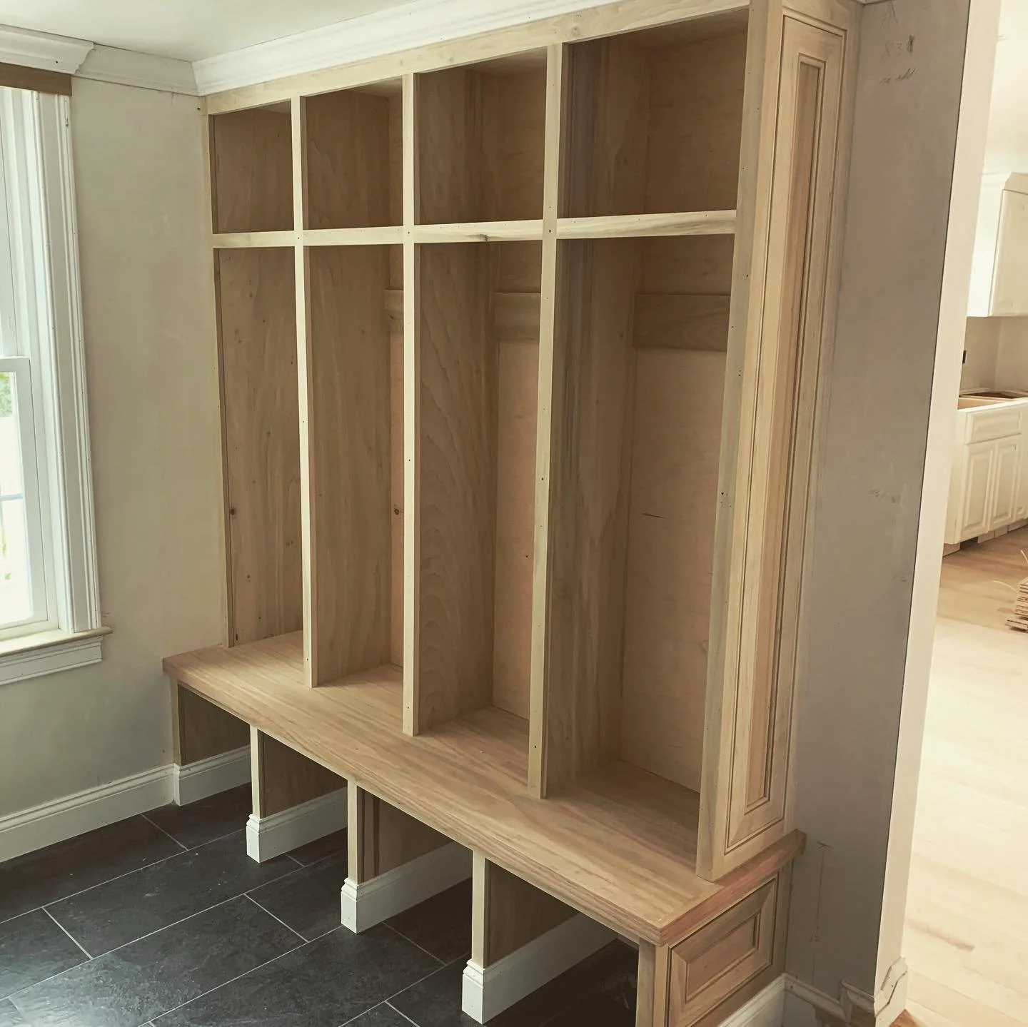 Unfinished wooden built-in shelving unit in a room with dark tile flooring and a window, showing vertical sections and a decorative panel on the side.