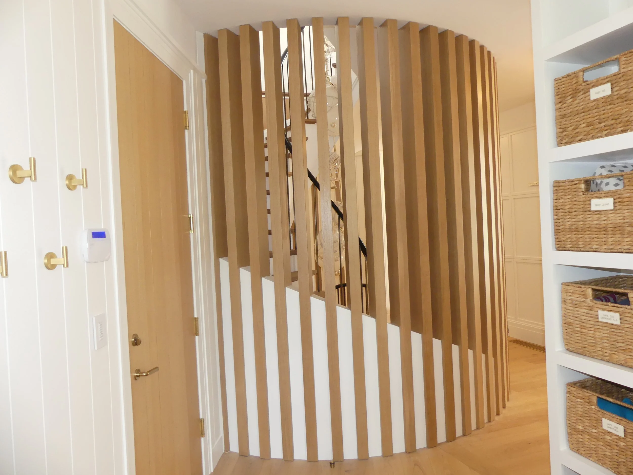 Interior view of a home featuring a curved wooden slat staircase with black handrails, a wooden door, and white wall-mounted storage baskets with labels on shelves.
