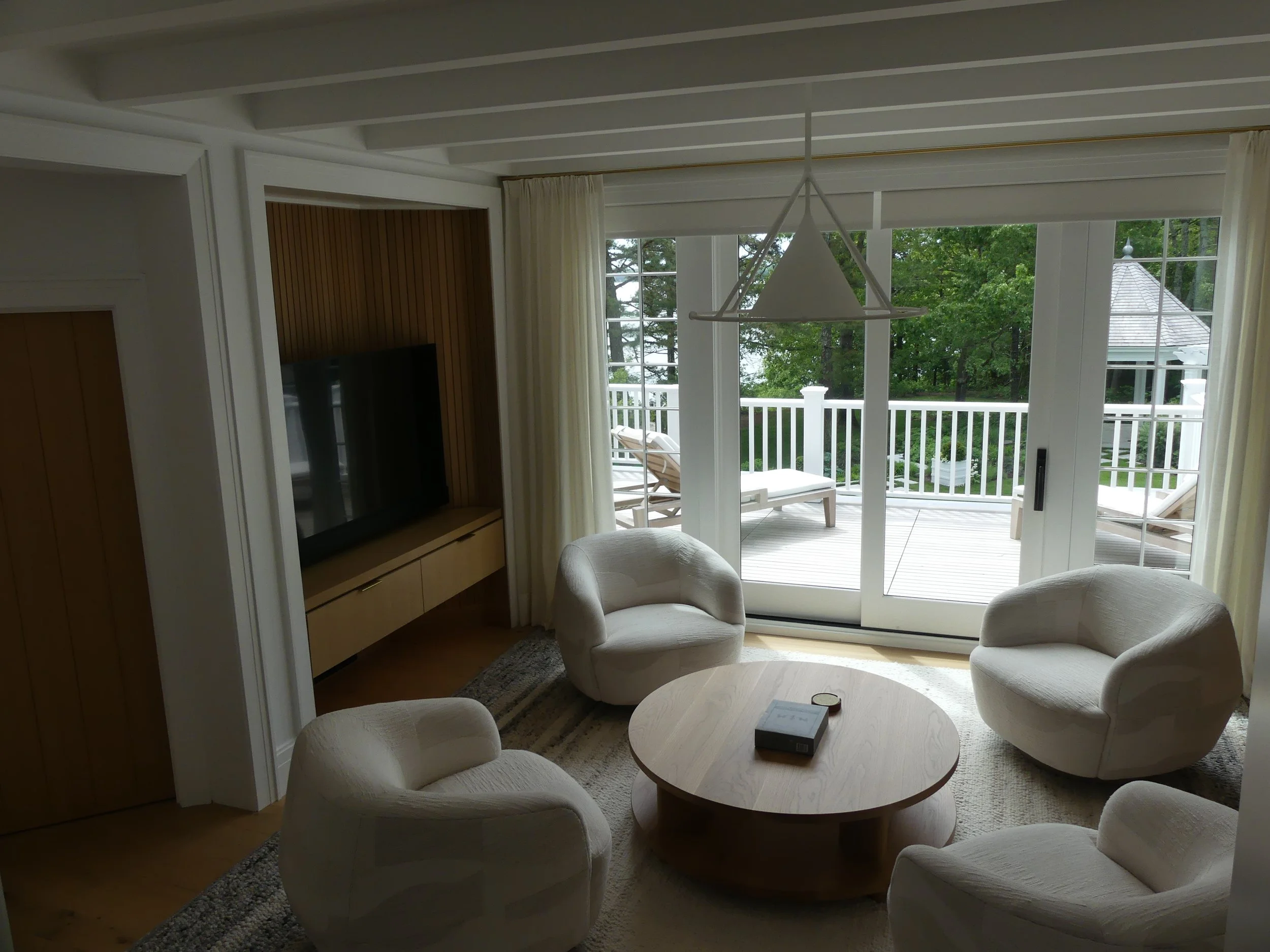 Living room with four white armchairs around a round wooden coffee table, large sliding glass doors leading to a patio with reclining chairs, TV on a wooden stand, and green trees outside.