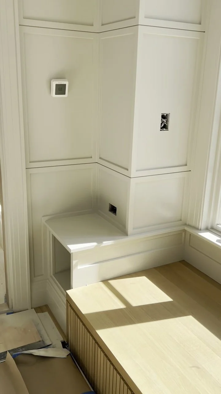 White wall with a thermostat, electrical outlets, and an empty space for switches, under construction, with a wooden bench and natural light from a nearby window.