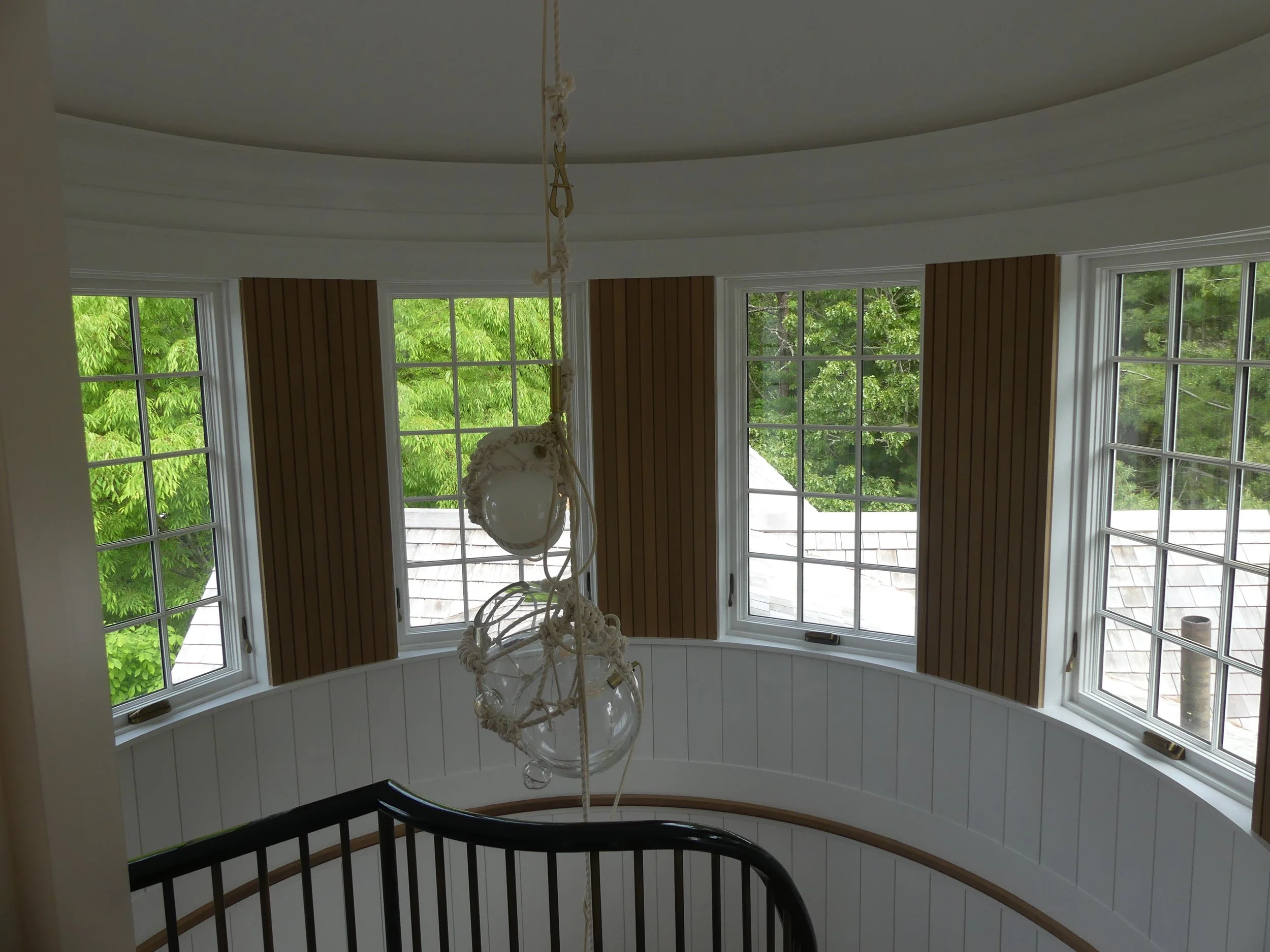 View of a curved wall with five windows and wooden shutters, overlooking green trees and a roof outside, with a black staircase and a hanging glass light fixture in the foreground.