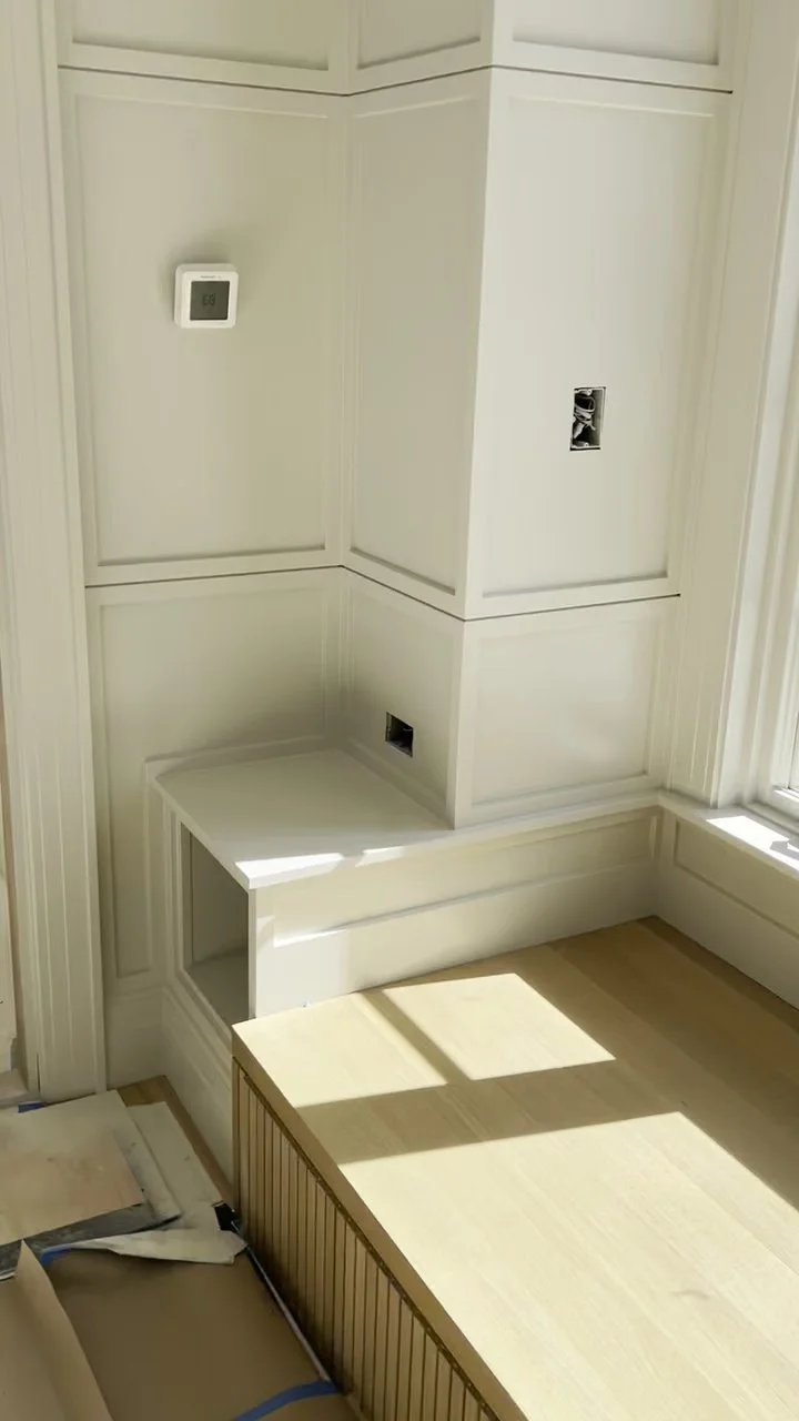 Partially built white wall with electrical outlets and a thermostat, next to a window with sunlight, and a wooden bench or platform in a room under construction.