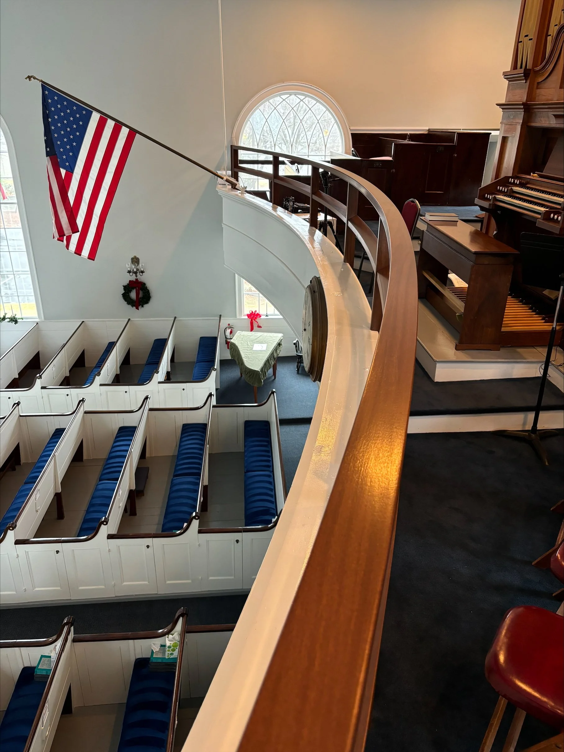 Interior view of a church or religious venue decorated for Christmas, featuring red, white, and blue American flags, a Christmas wreath, and empty wooden pews with blue cushions, seen from an upper balcony overlooking the nave, with organs on the rig