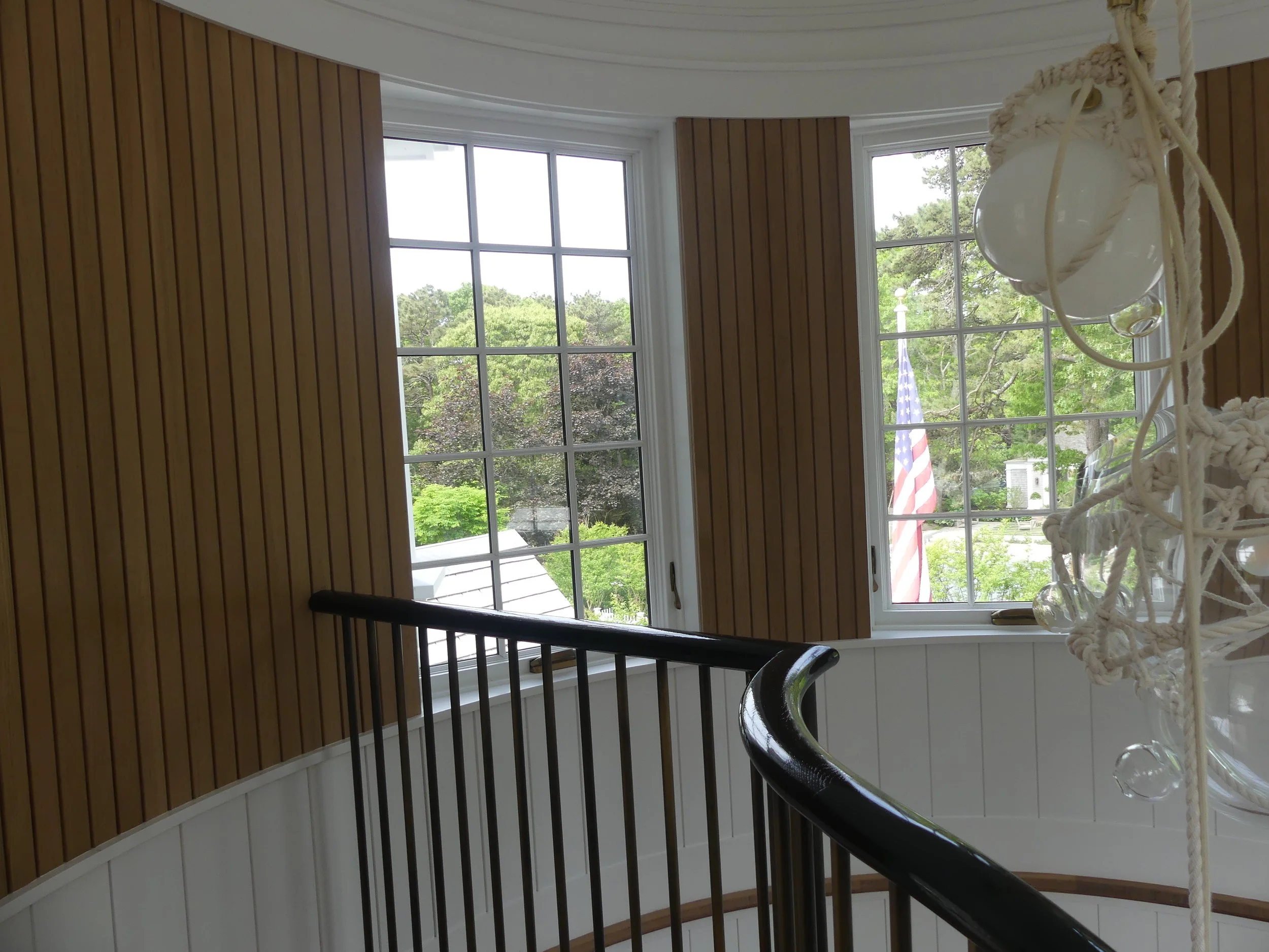 Interior of a house featuring two large windows, a curved staircase with a black handrail, and a decorative light fixture with exposed bulbs and rope accents
