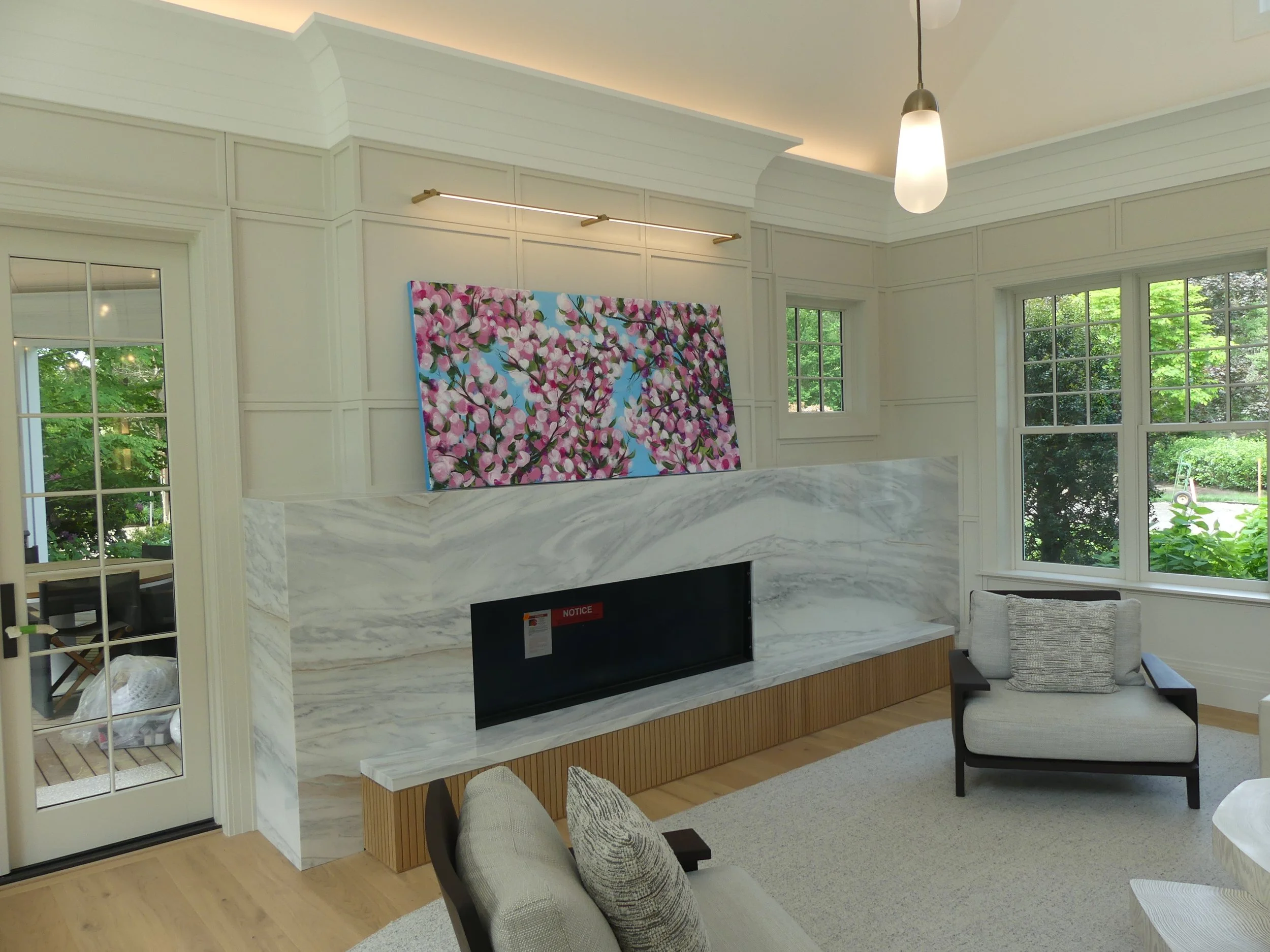 Living room with a marble fireplace, large windows showing greenery outside, a painting of pink blossoms above the fireplace, modern ceiling lighting, and a white armchair with a gray cushion.