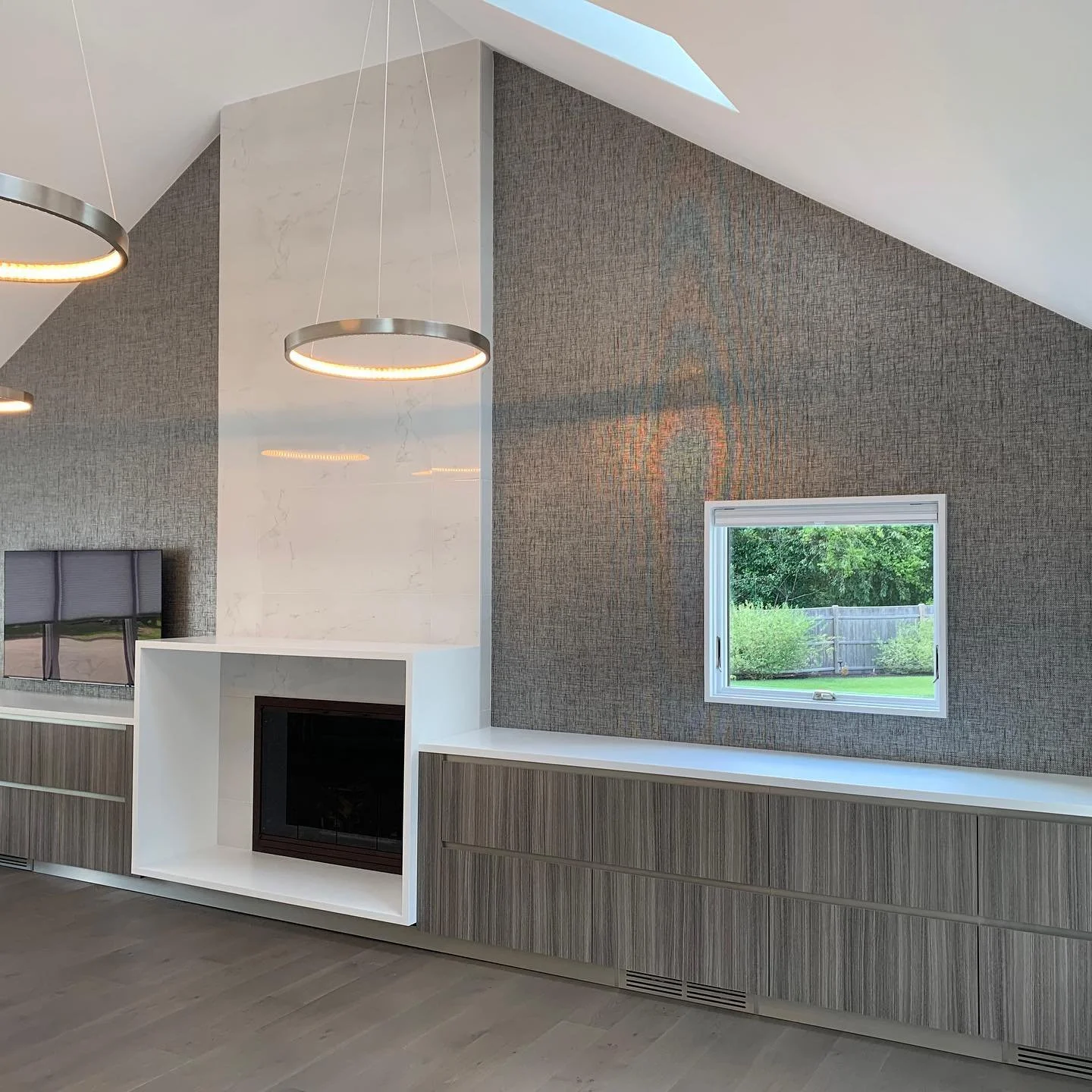 Modern living room with built-in white fireplace, textured gray wall, and large wall-mounted TV. Ceiling with skylight and circular pendant lights.