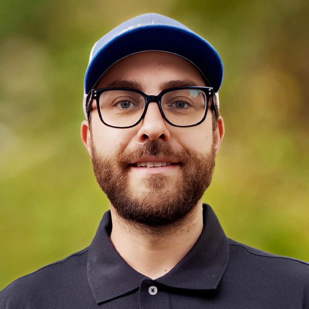 A man wearing glasses, a black polo shirt, and a blue cap, smiling outdoors with a blurred green background.