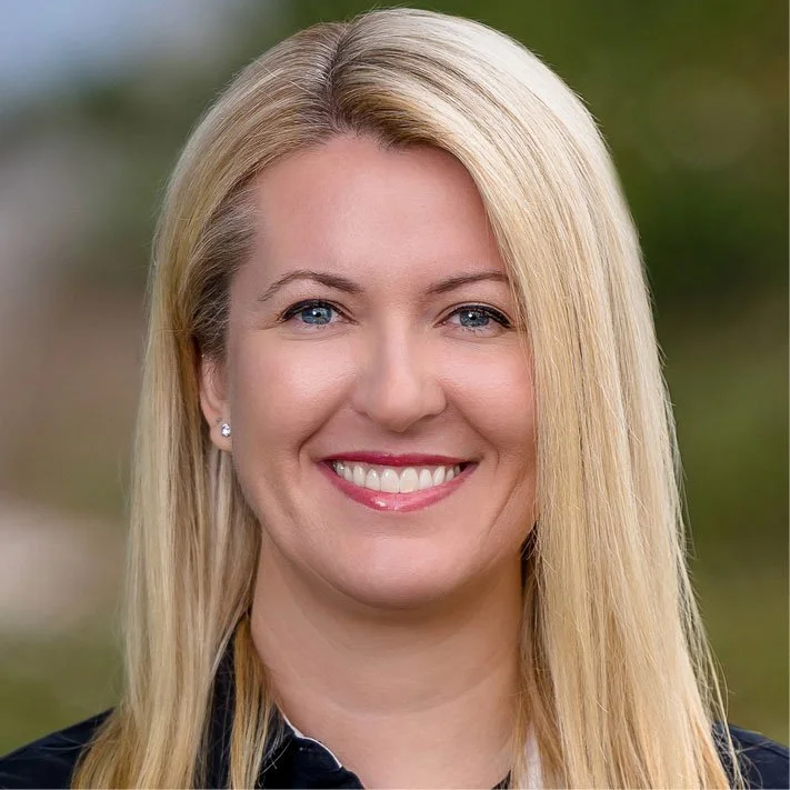 A woman with long blonde hair smiling outdoors with a blurred green background.