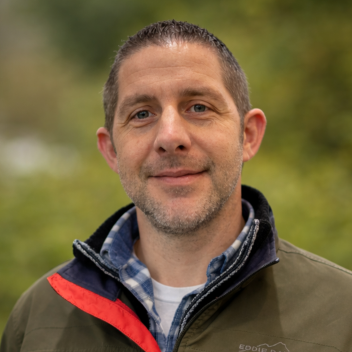 Portrait of a middle-aged man with short dark hair and slight beard, wearing a green jacket over a plaid shirt, standing outdoors with blurred green trees in background.