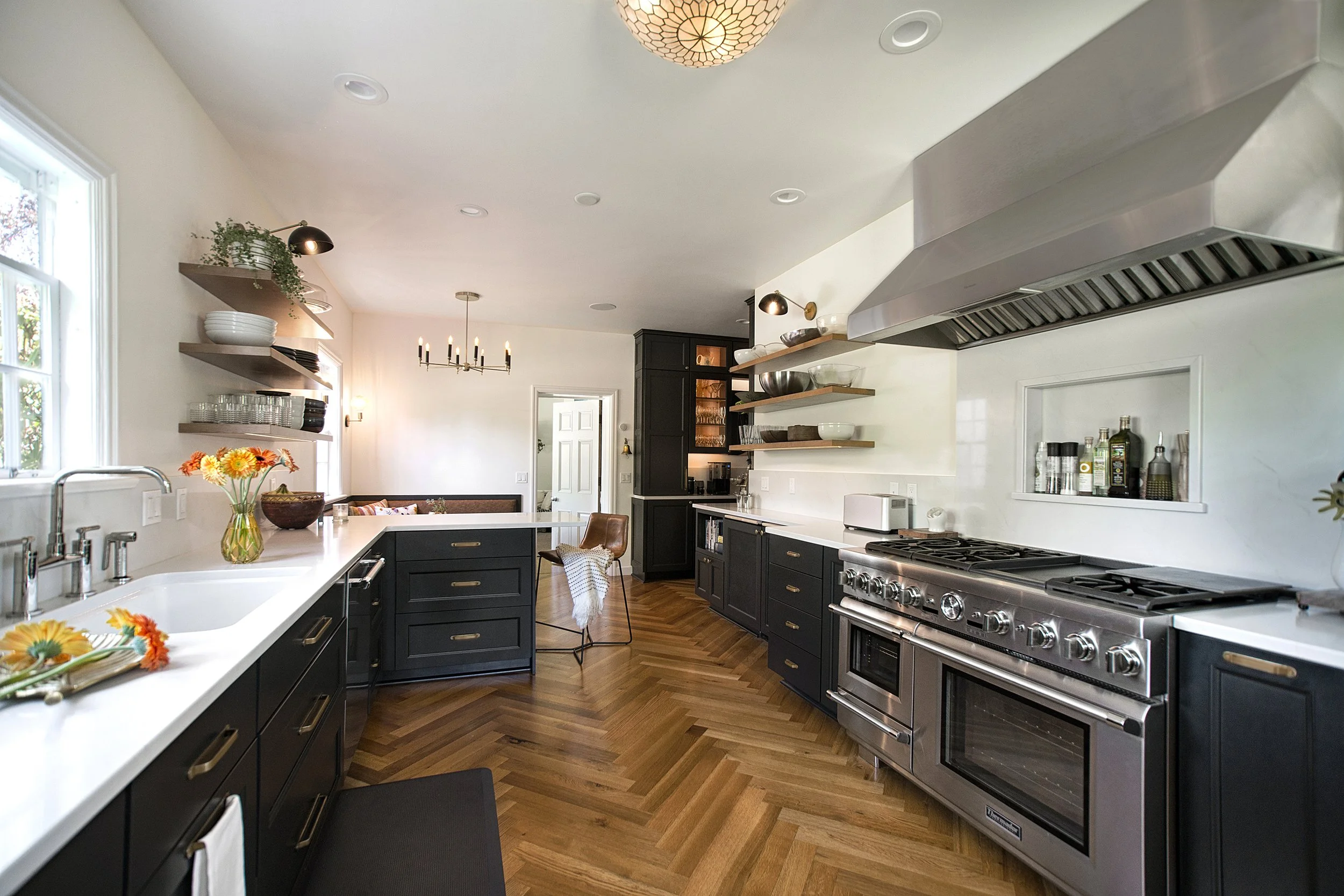 Modern kitchen with black cabinets, stainless steel appliances, open wooden shelves, white countertops, and a herringbone wood floor.