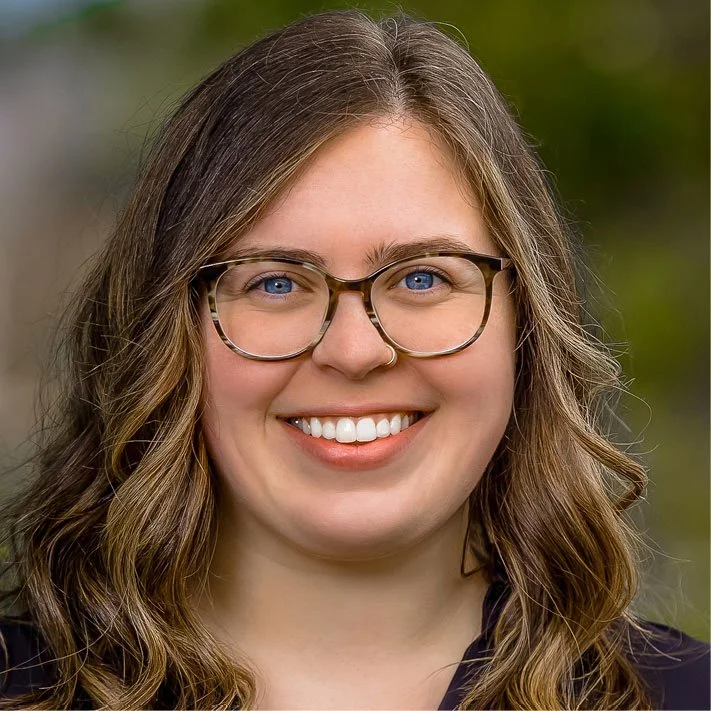 Close-up portrait of a young woman with wavy brown hair, glasses, and a nose ring, smiling outdoors with a blurred green background.
