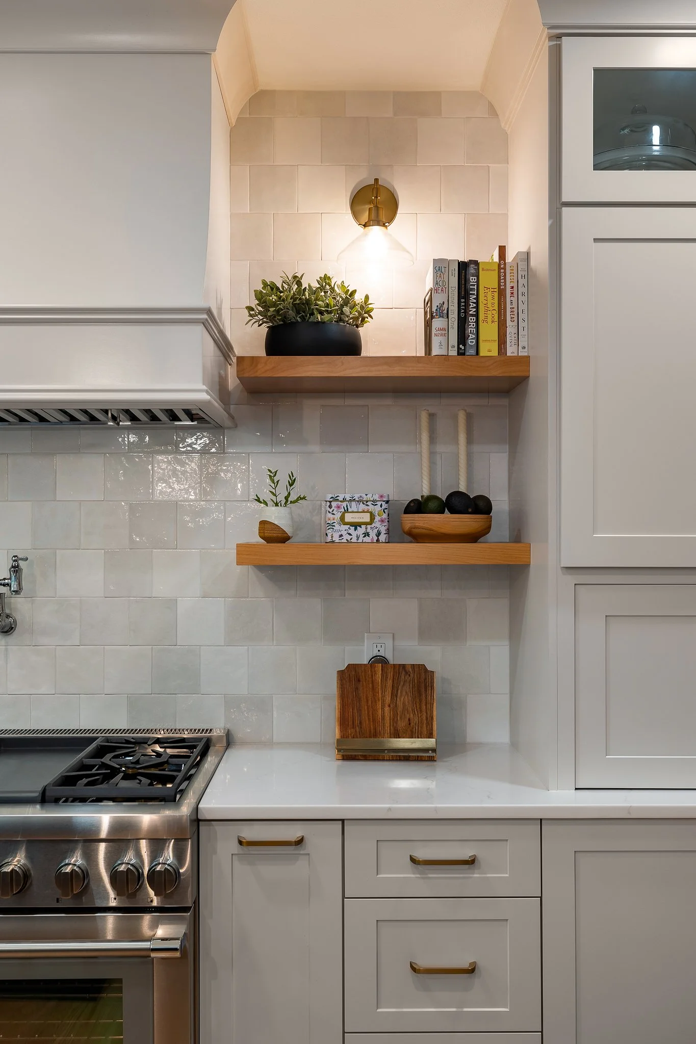 Kitchen with white cabinets, beige tile backsplash, stainless steel stove, and wooden open shelves holding potted plants, books, and decorative items, with a wall light above.