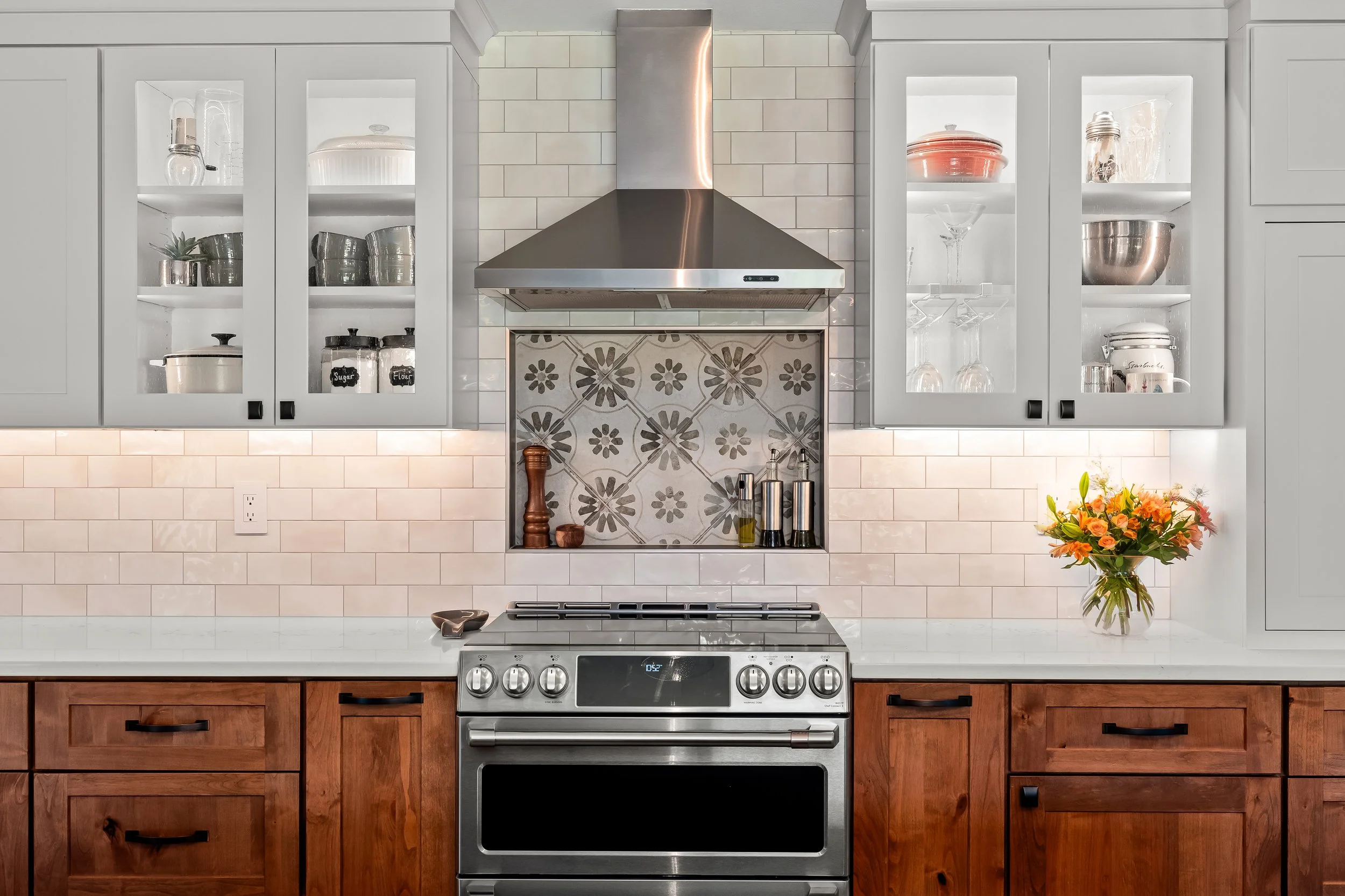 Kitchen with white cabinets, a stainless steel stove, and wooden lower cabinets. Glass-front upper cabinets contain various dishes and glassware. A bouquet of flowers is on the right counter, and a spoon rest is on the left of the stove. The backdrop