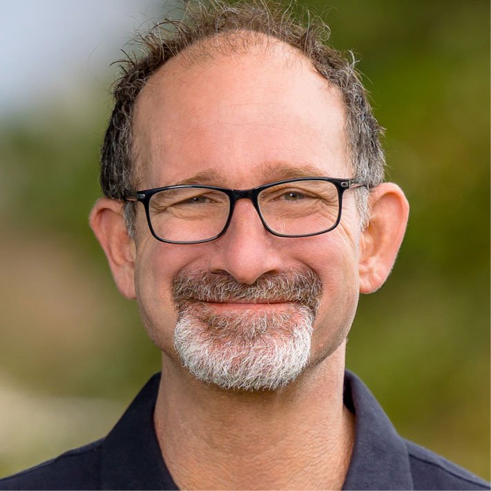 Close-up of a middle-aged man with glasses, graying hair, and a beard, smiling outdoors with a blurred green background.