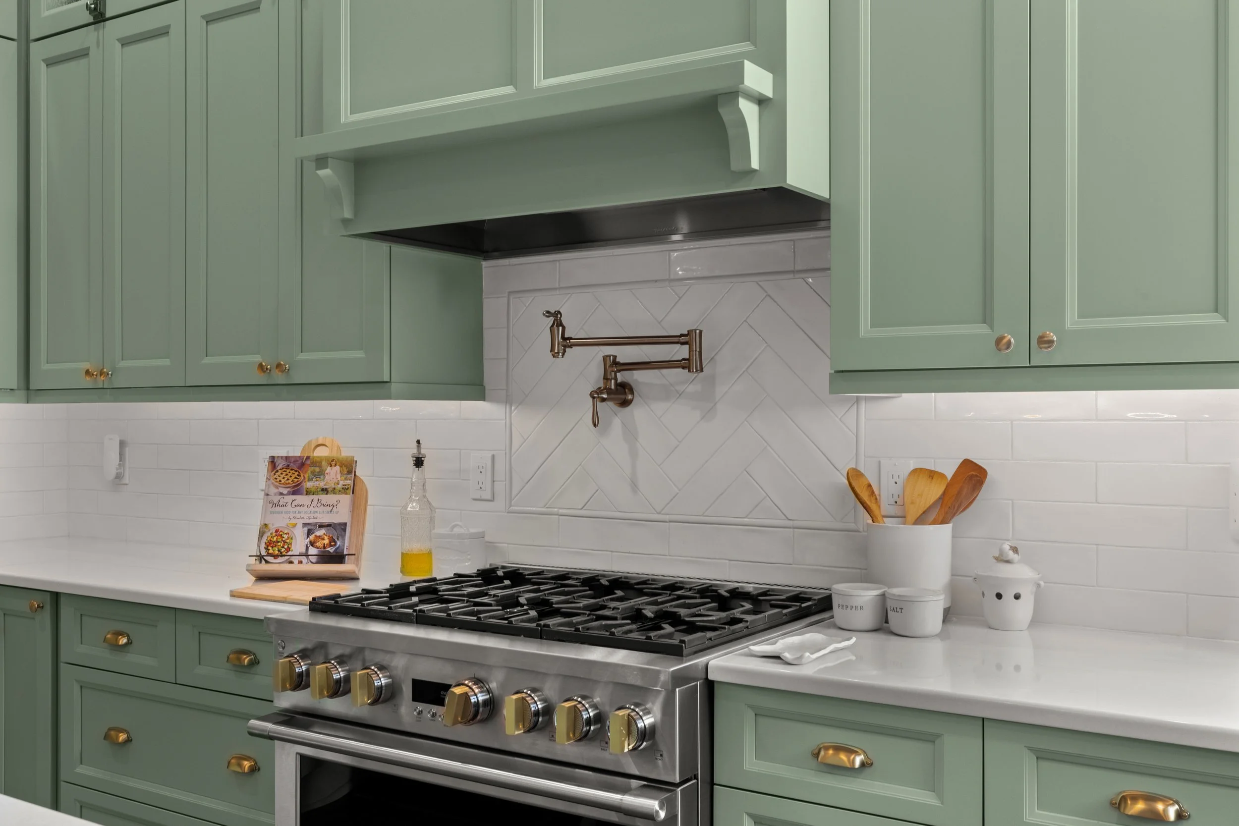 Kitchen with mint green cabinets, white subway tile backsplash, and a stainless steel gas stove with gold knobs. Countertop has a cookbook stand, oil dispenser, and storage jars. Gold cabinet hardware and a brass pot filler above the stove.