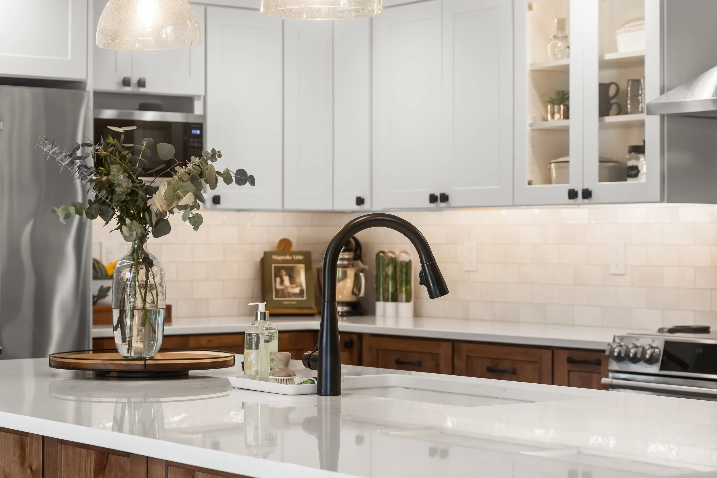 Modern kitchen with white cabinets, a black faucet, and a marble countertop. Decor includes a vase with greenery, a soap dispenser, and a tray with small items. Light fixtures hang from the ceiling.
