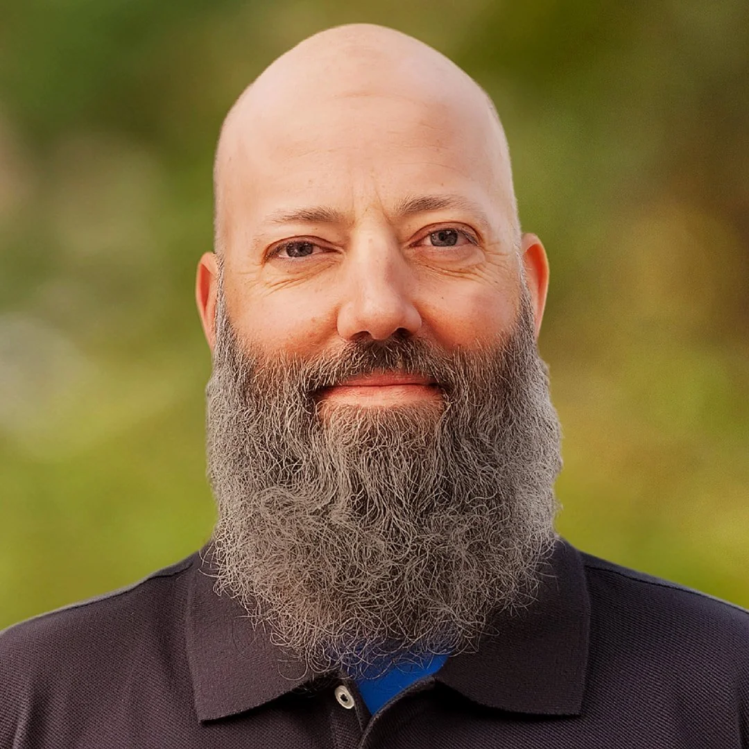 Headshot of a bald man with a long gray beard, wearing a dark collared shirt, smiling outdoors with a blurred green background.