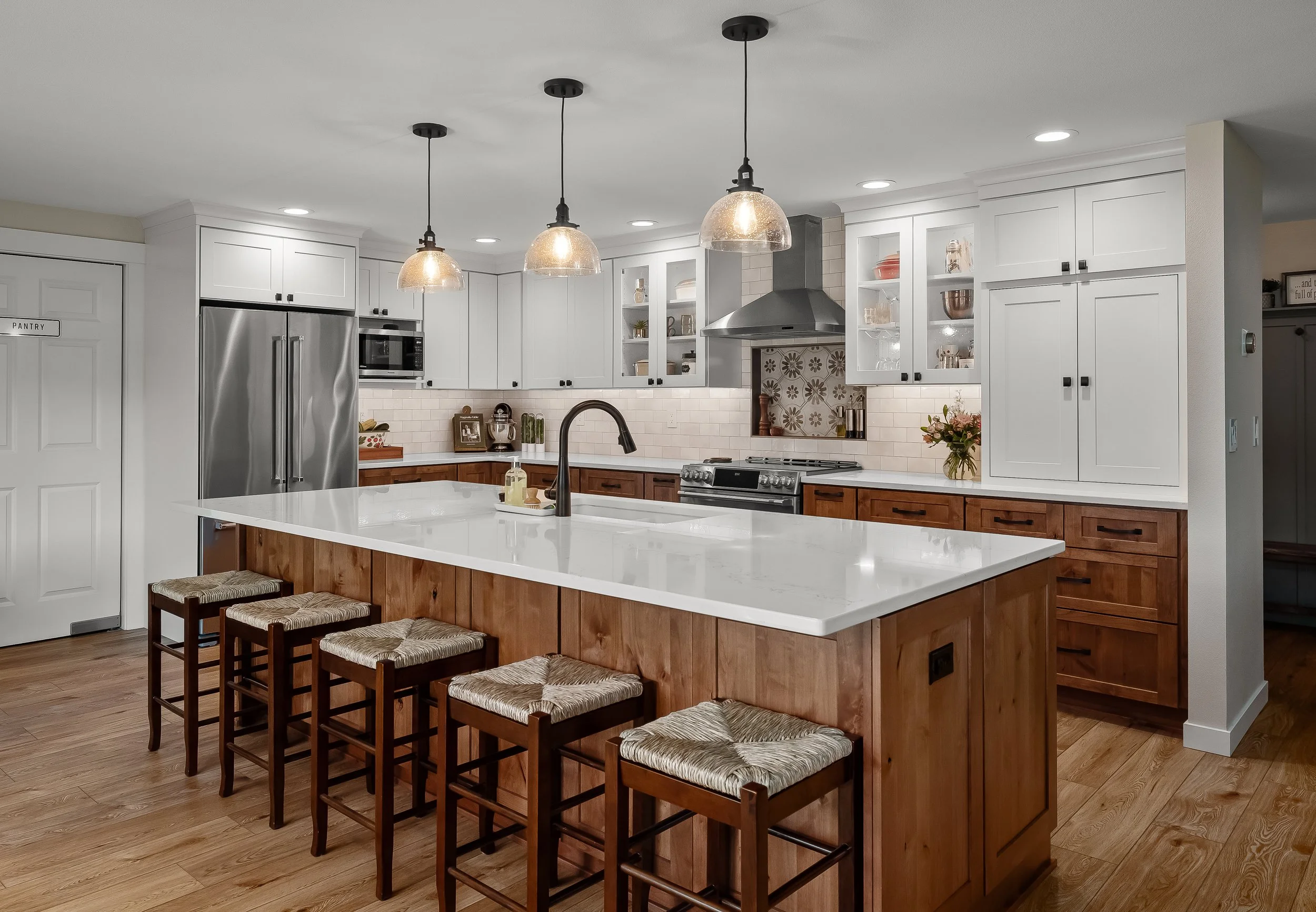 Kitchen with white cabinets, stainless steel refrigerator, and a large island with a white countertop and wooden base. Four wooden chairs with woven seats are placed at the island. There are pendant lights hanging above the island.
