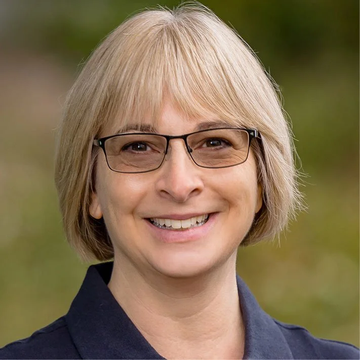 A smiling woman with blonde hair, wearing glasses and a dark shirt, outdoors with blurred greenery in the background.