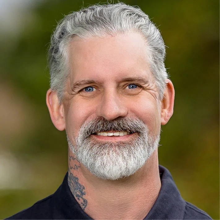 Close-up of a smiling middle-aged man with gray hair and beard, blue eyes, wearing a dark shirt, outdoors with blurred green background.