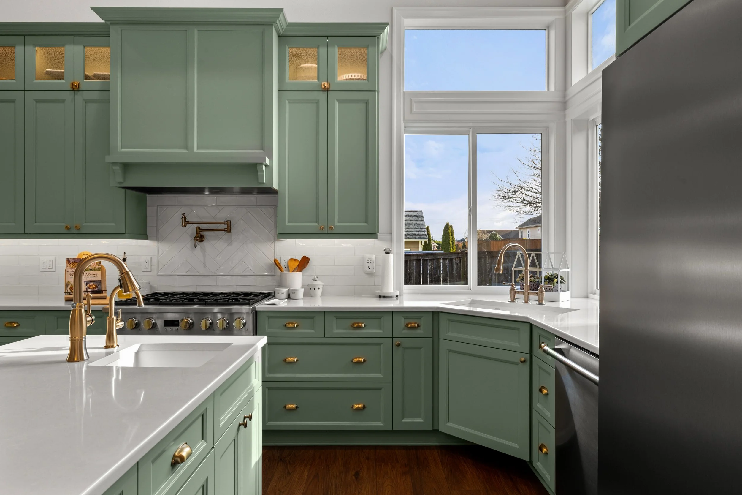 Modern kitchen with green cabinets, white countertops, and a view of a backyard through large windows.