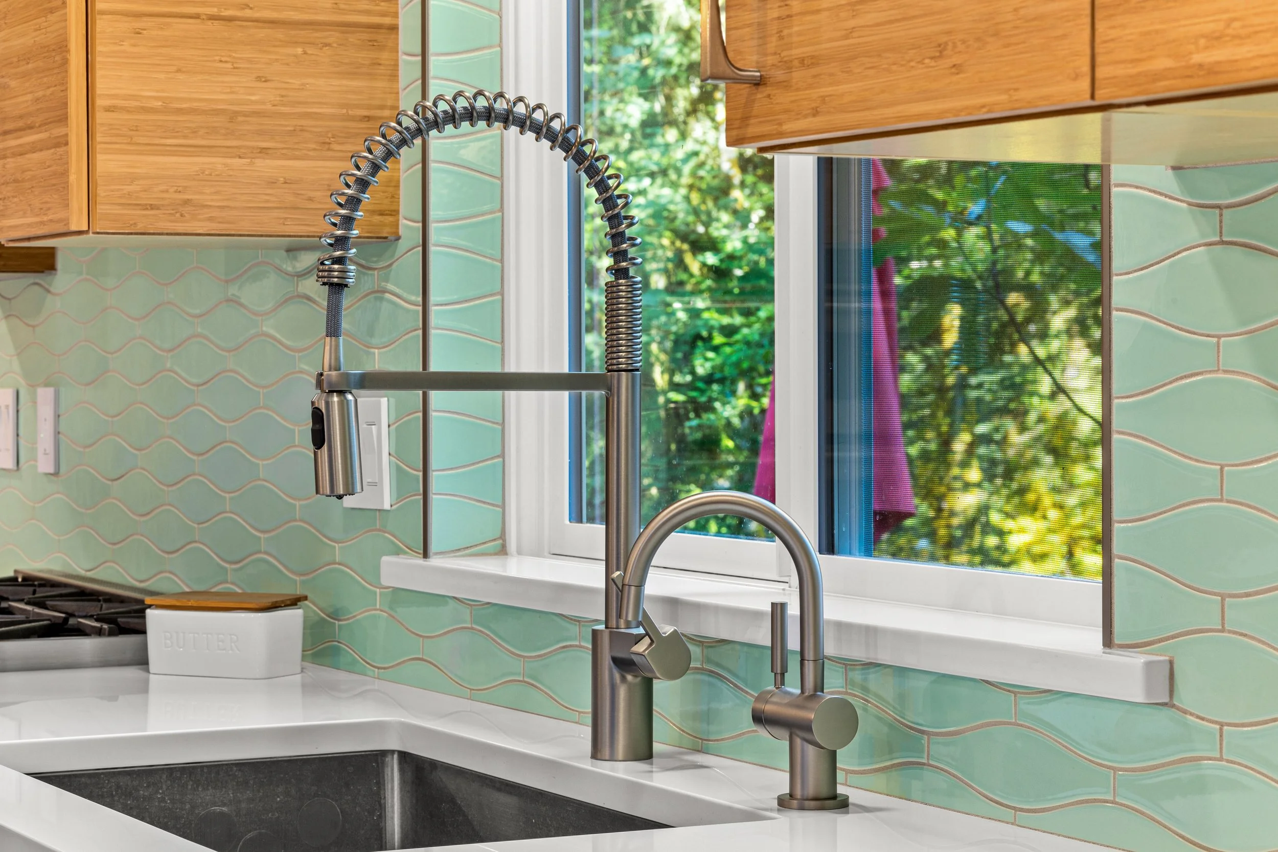 Modern kitchen sink with dual faucets, a window showing greenery outside, and a green tiled backsplash.