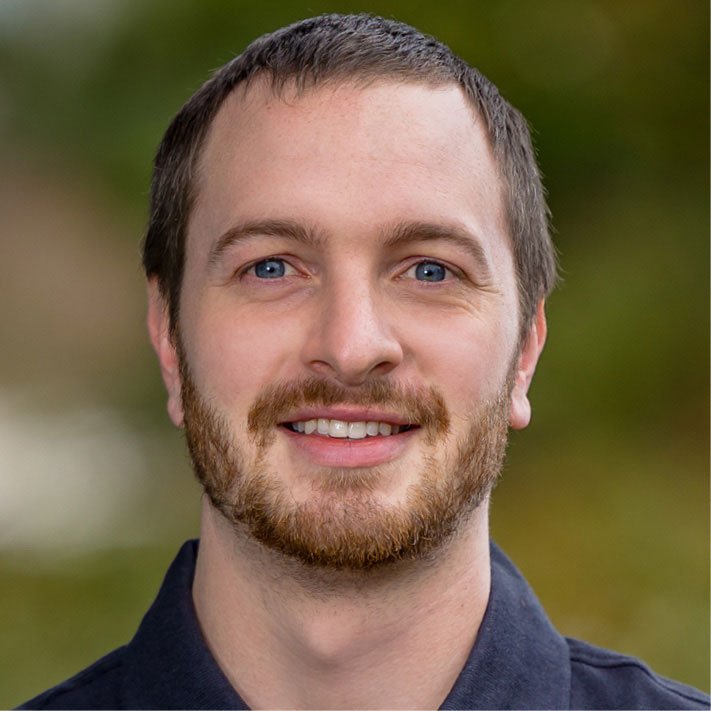 A young man with blue eyes, light brown hair, and a beard smiling outdoors against a blurred green background.