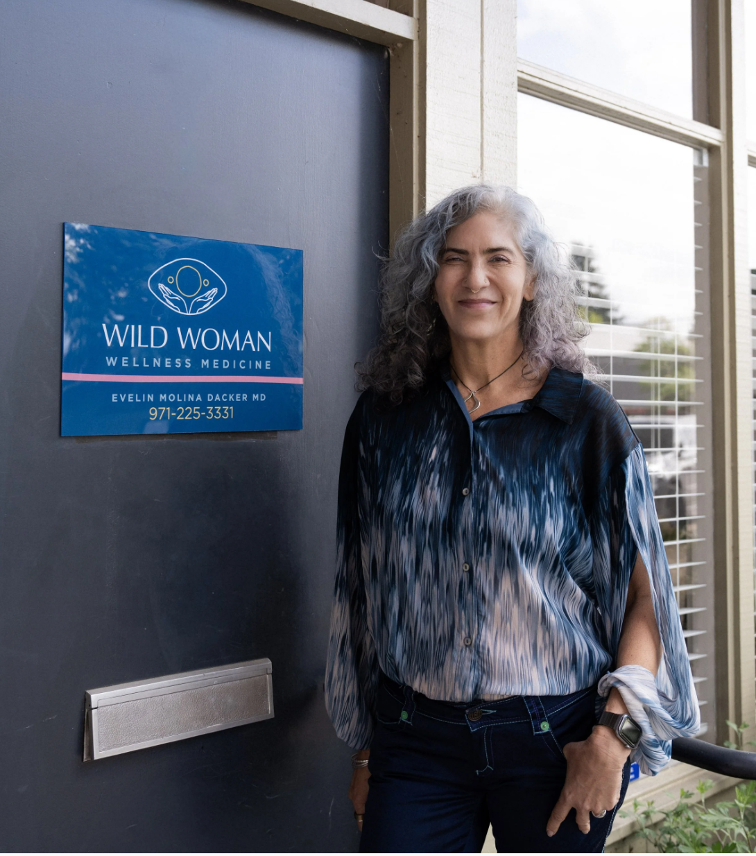 Evelin Molina Dacker M.D, with gray curly hair standing outside near a door with a sign that says Wild Woman Wellness Medicine, with contact information, smiling slightly, wearing a multicolored blouse and a watch.