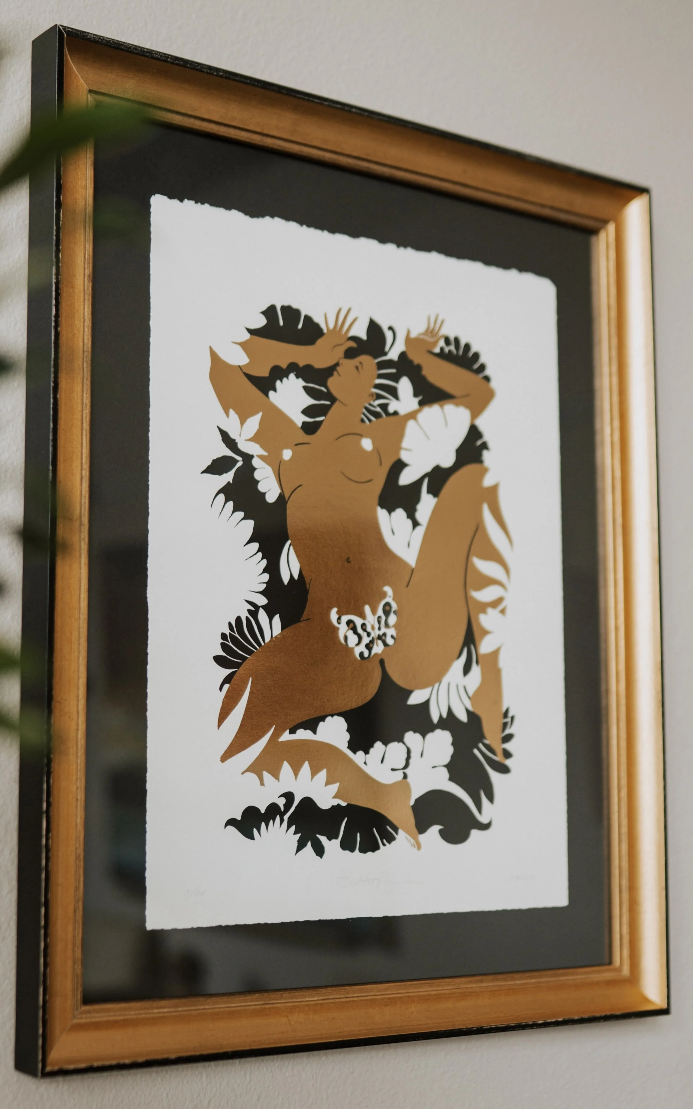 Framed artwork of a stylized female figure with long hair, adorned with flowers and leaves, in brown, black, and white colors, mounted on a wall.