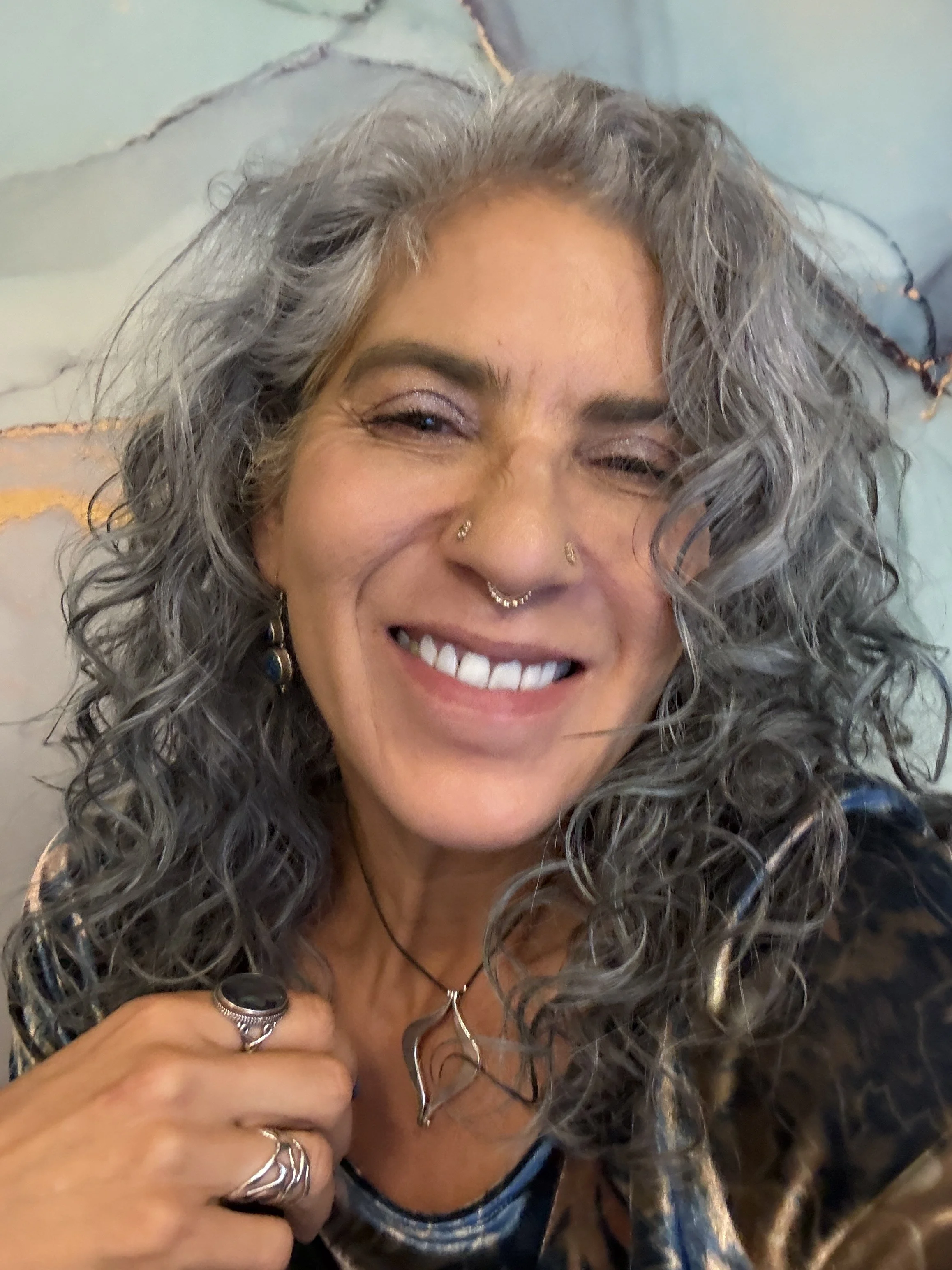 Evelin Molina Dacker M.D, with curly gray hair smiling, wearing rings and necklaces, with a blue and black shirt.