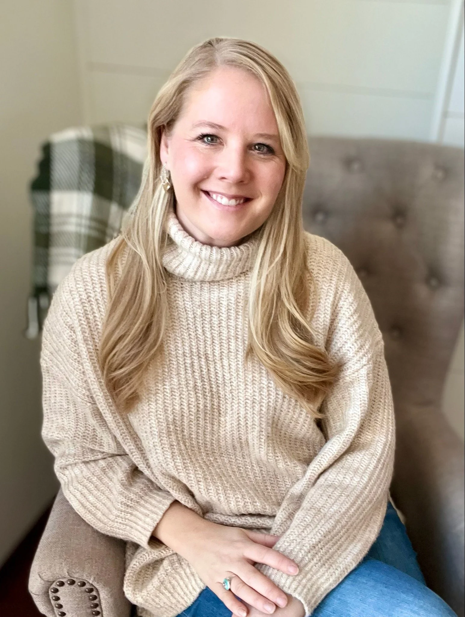 A smiling woman with long blonde hair wearing a beige turtleneck sweater and sitting on a beige chair with a button back in a cozy indoor setting.