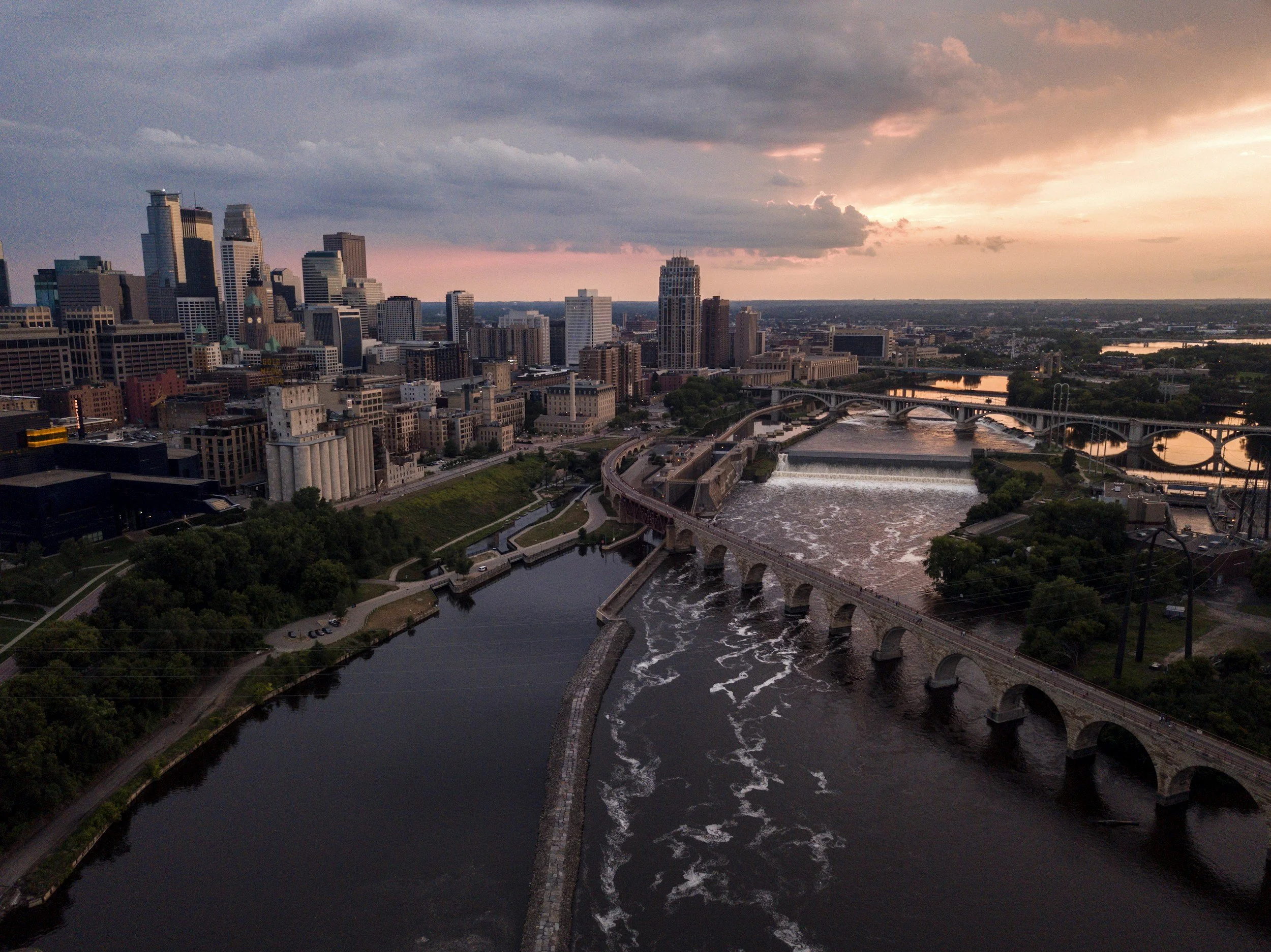 An aerial view of downtown Minneapolis at sunset, showing skyscrapers, the Mississippi River with dams and bridges, and a partly cloudy sky with pink and orange hues.