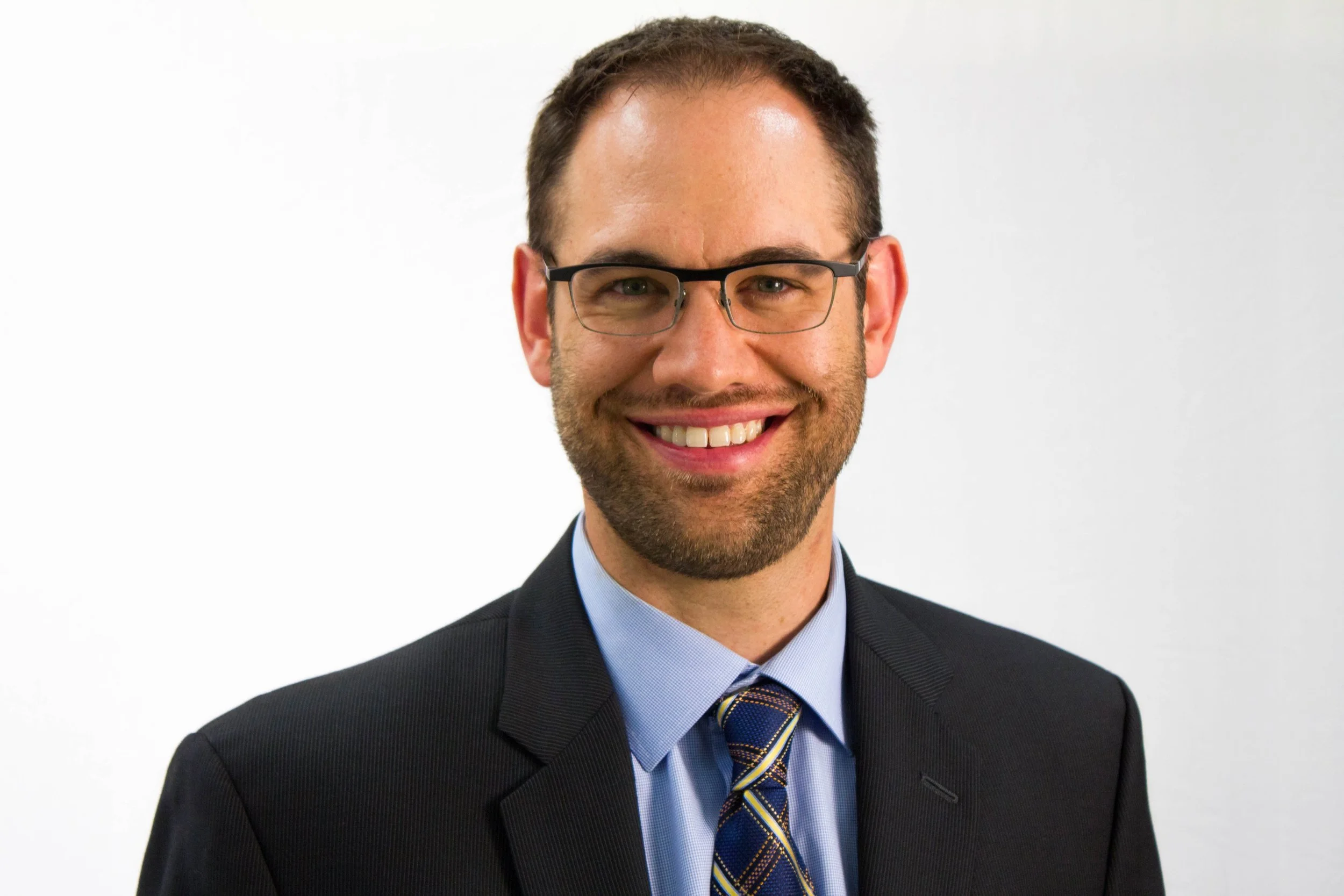 Headshot of a smiling man with glasses, dressed in a suit and tie, against a plain white background.