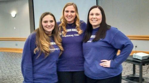 Three young women standing together indoors, smiling at the camera, wearing matching blue sweatshirts.