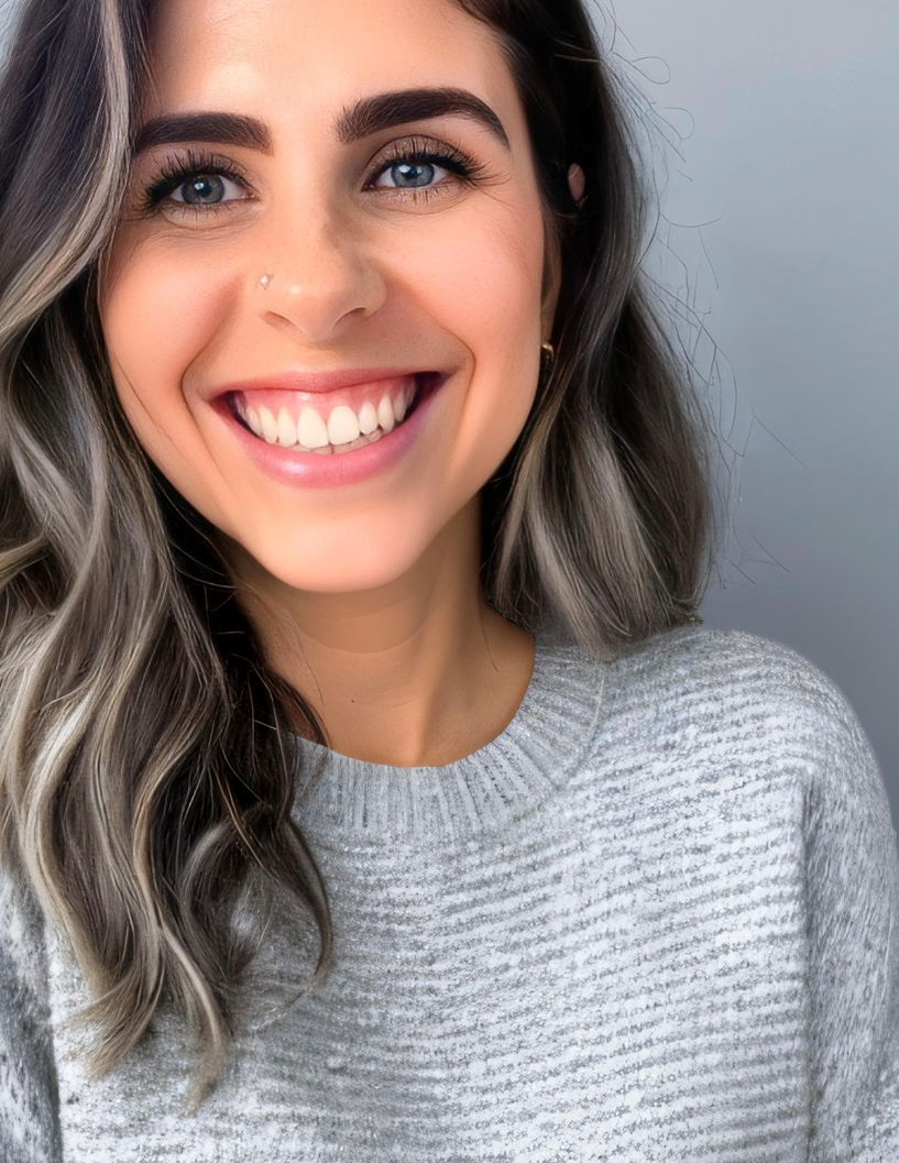 Close-up of a young woman smiling with blue eyes, dark eyebrows, and wavy, blonde and dark brown hair, wearing a gray knit sweater.