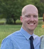 A smiling man with a shaved head wearing a blue shirt and tie, outdoors with trees in the background.