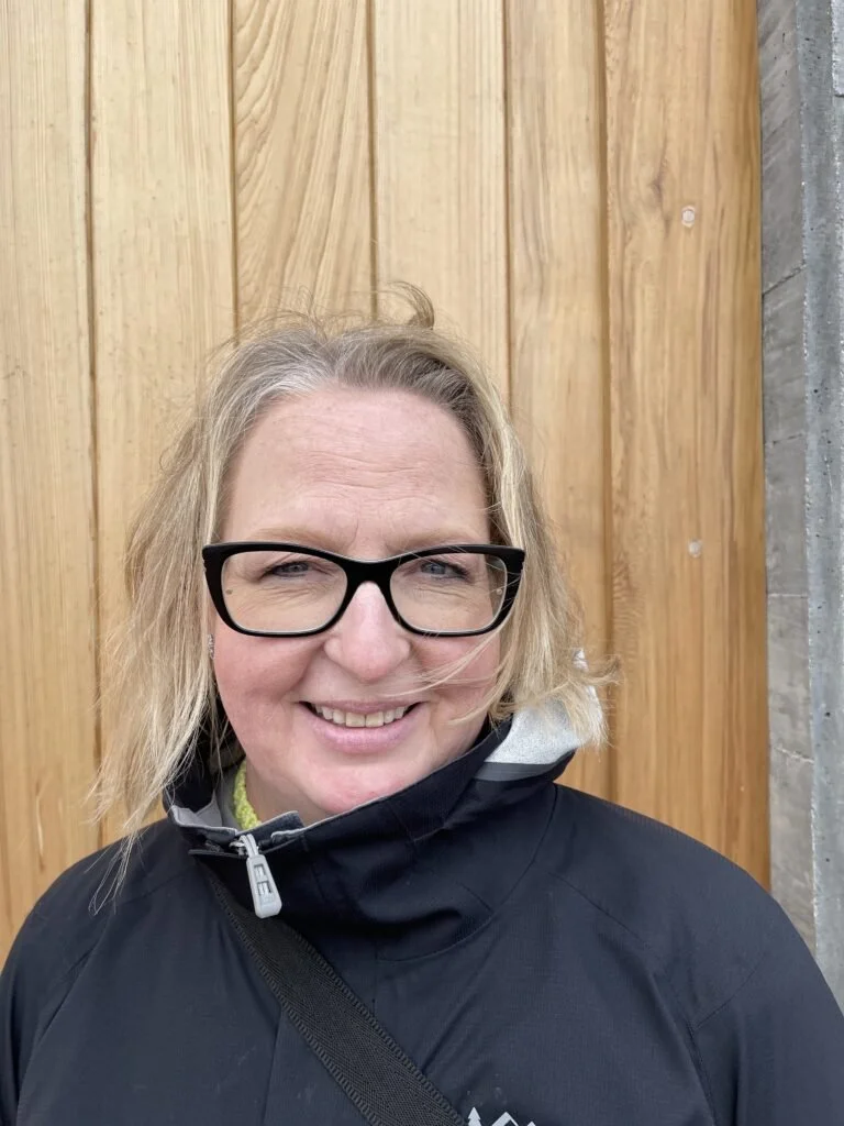 Woman with blonde hair, glasses, and a black jacket standing in front of a wooden wall.