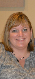 Smiling woman with shoulder-length blonde hair, wearing a patterned blouse and a black necklace.