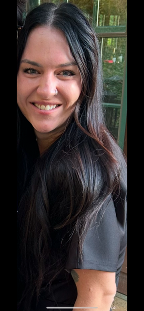 A woman with long dark hair smiling indoors near a window with trees outside.