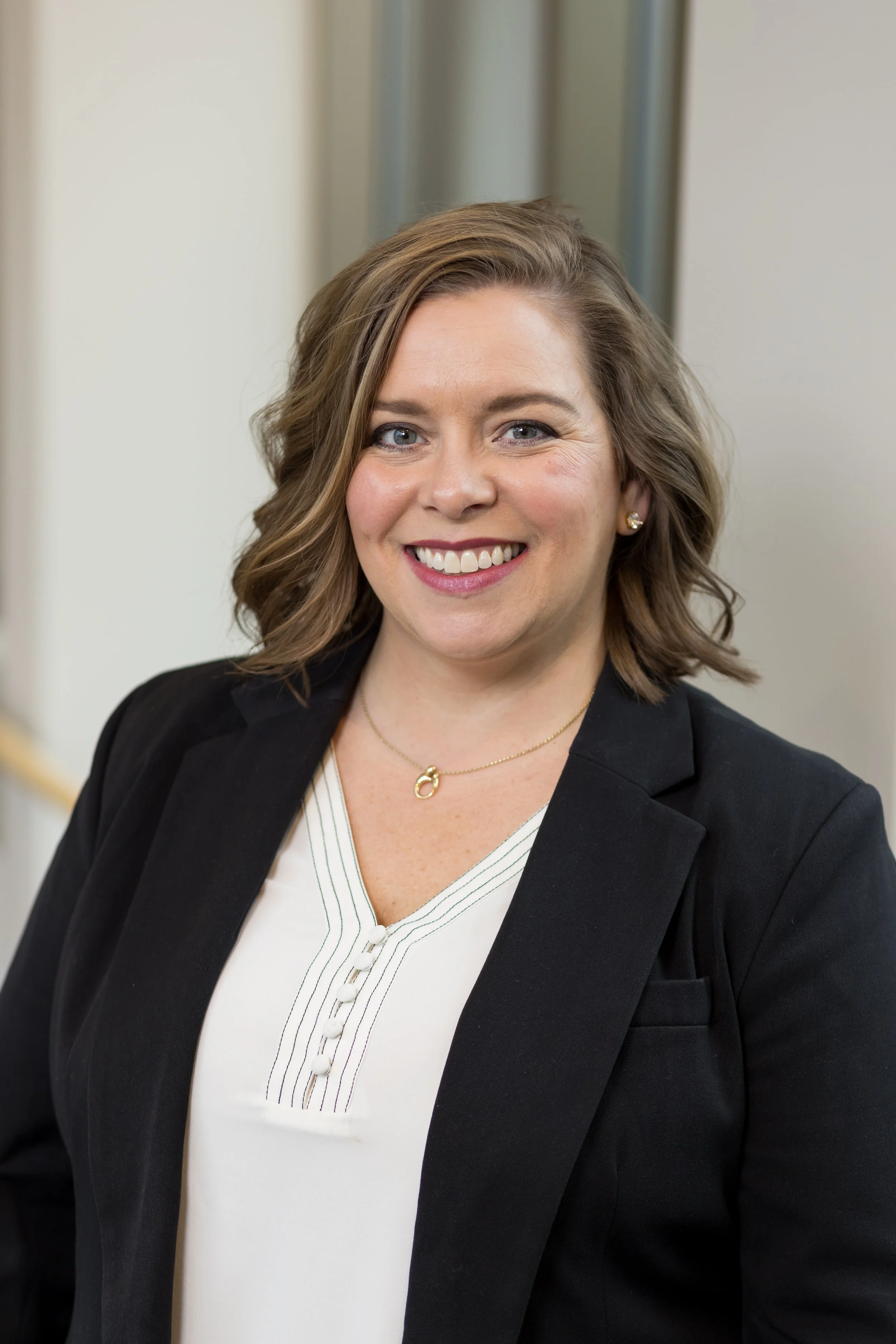 A woman with shoulder-length wavy brown hair, wearing a black blazer over a white blouse, smiling at the camera.