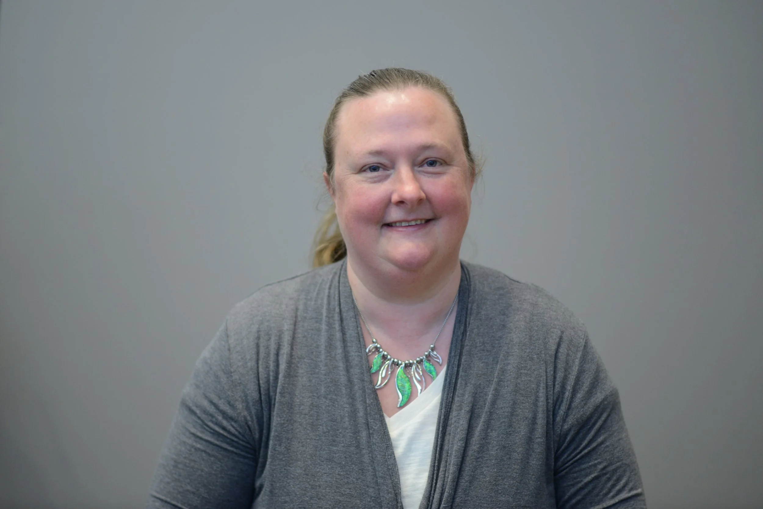 A woman with blonde hair tied back, wearing a gray cardigan and a colorful necklace, smiling at the camera against a plain gray background.