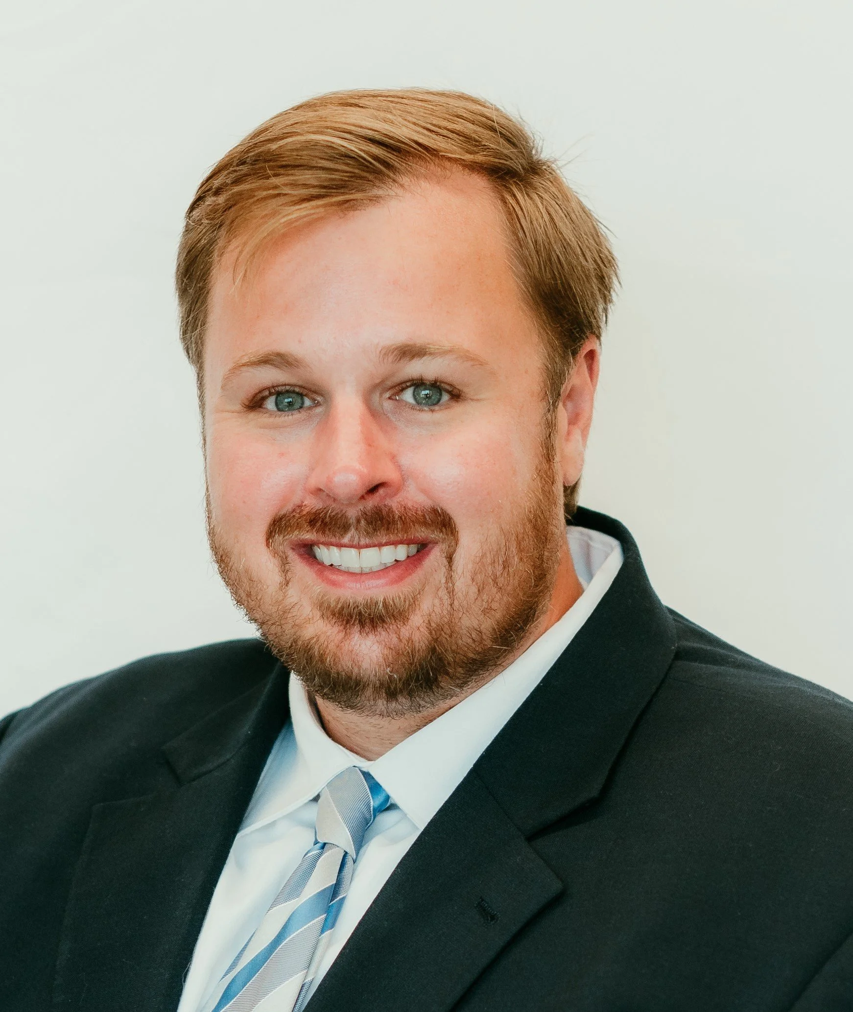 Headshot of a smiling man with light brown hair, beard, and blue eyes wearing a black suit and striped tie.