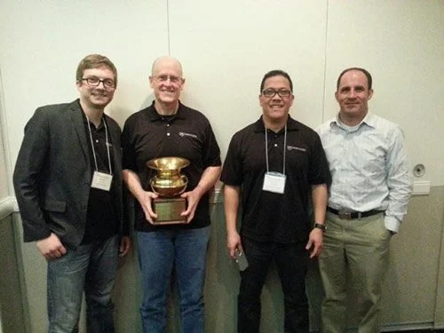 Four men standing side by side indoors, one holding a gold trophy, all smiling.