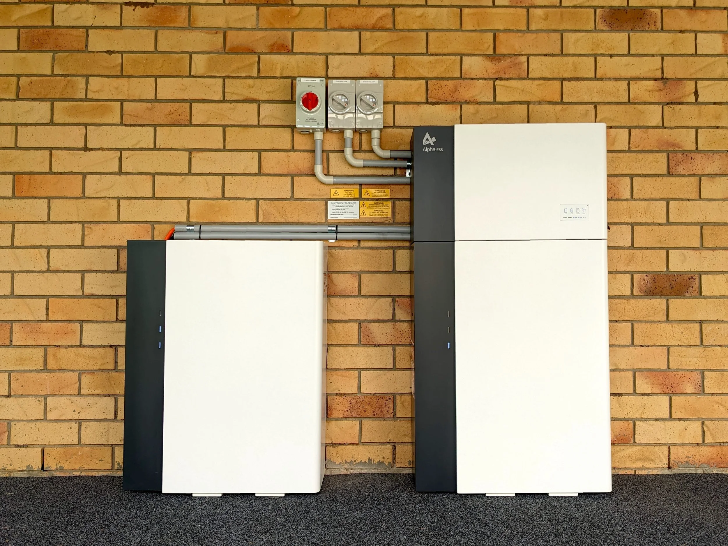 Home solar battery system mounted on exterior brick wall with inverter and electrical connections for residential solar power storage.