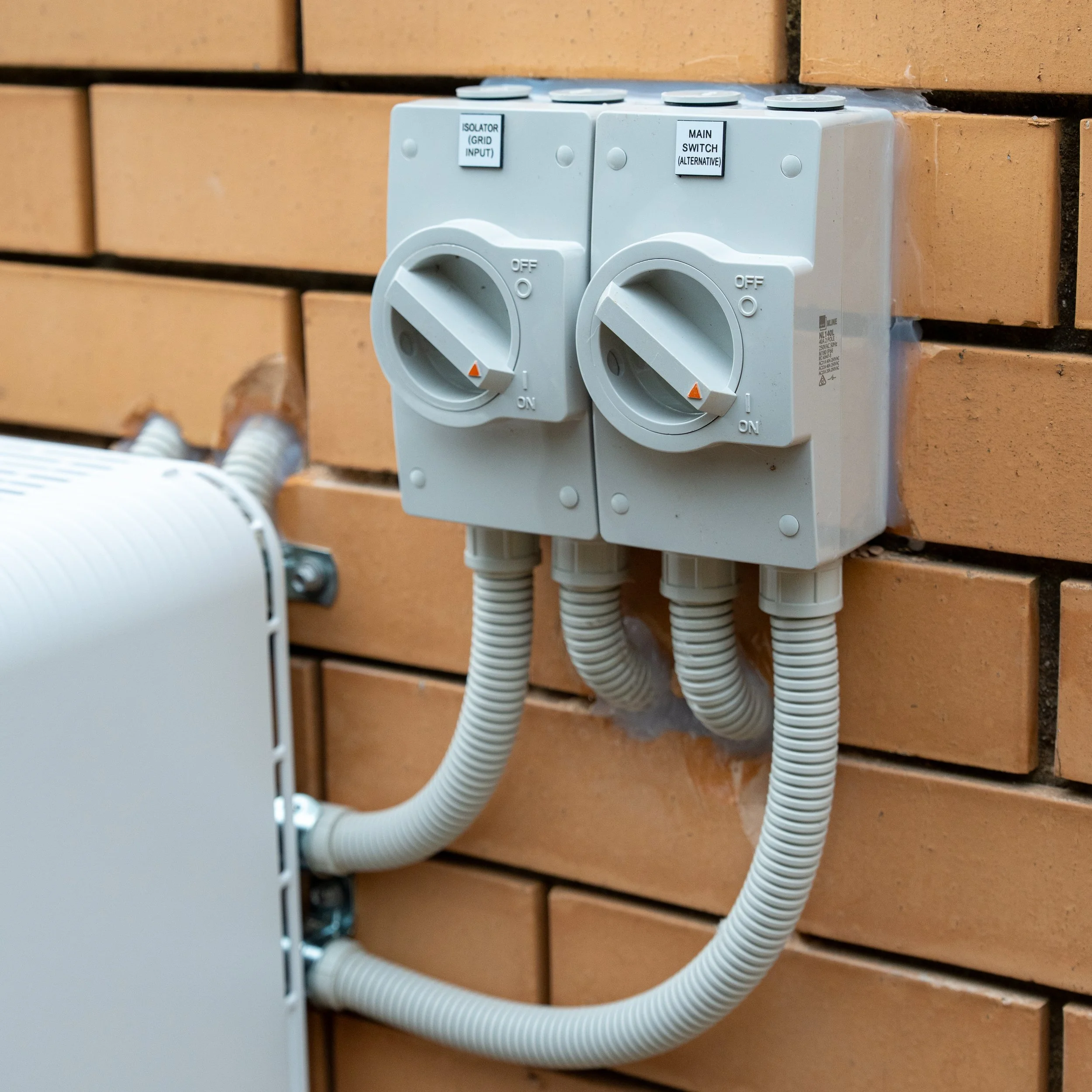 Electrical switch box with two switches and labeled buttons, mounted on a brick wall, with electrical conduit pipes connected.