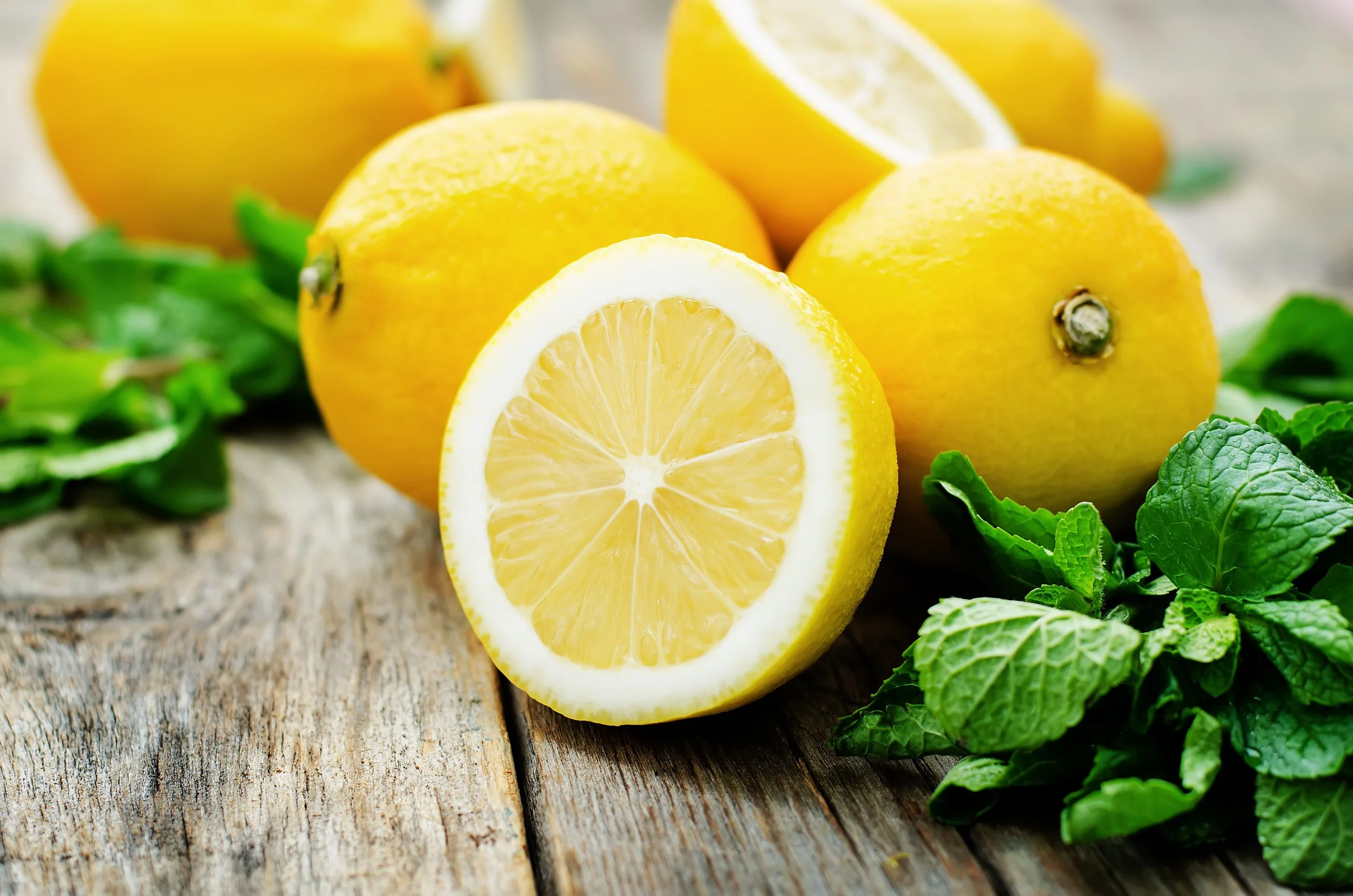 Fresh lemons, some sliced and some whole, with green mint leaves on a rustic wooden surface.
