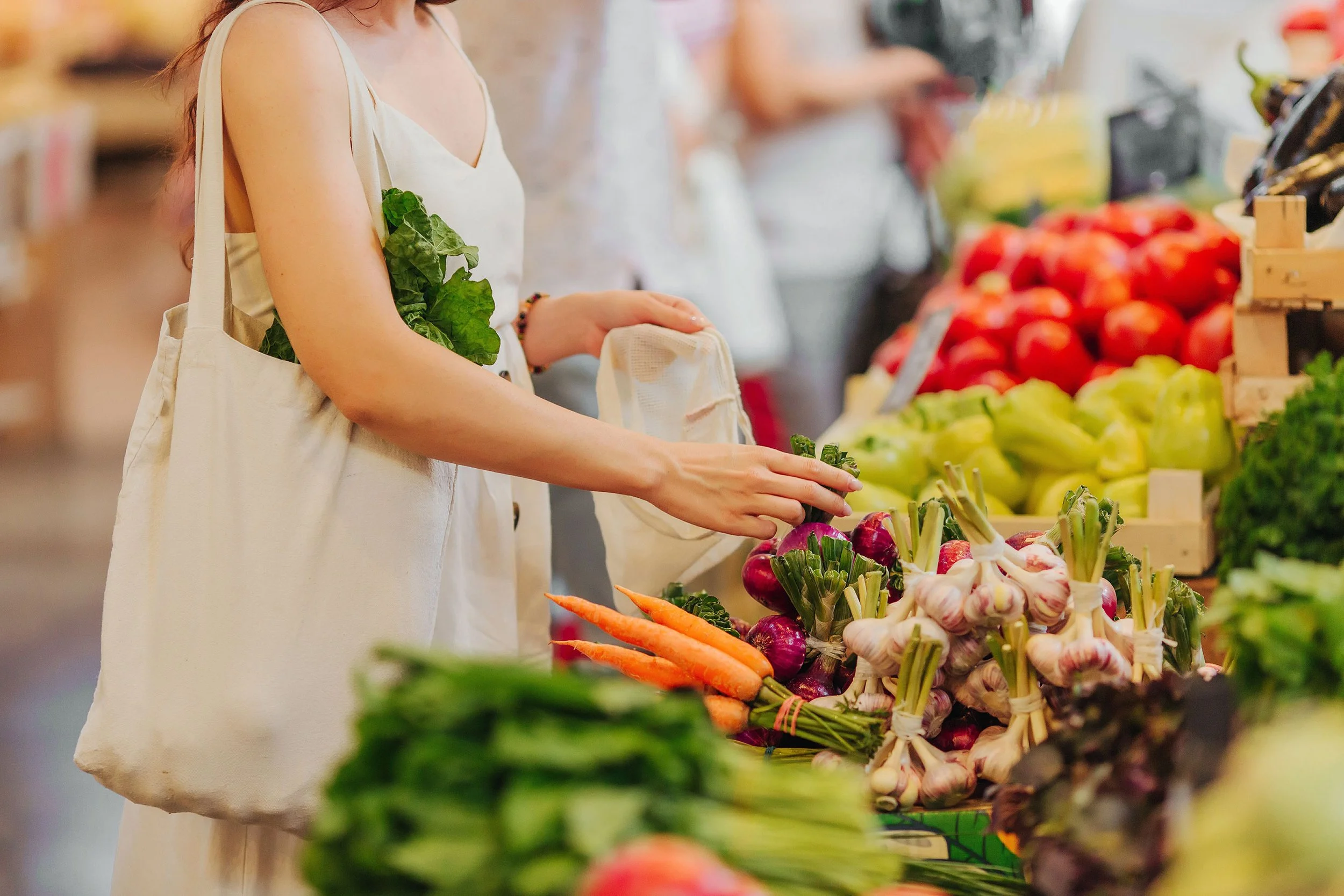 A woman shopping at a grocery store produces section, holding a bag and selecting vegetables, with various fresh produce like carrots, radishes, and greens displayed on the shelves.