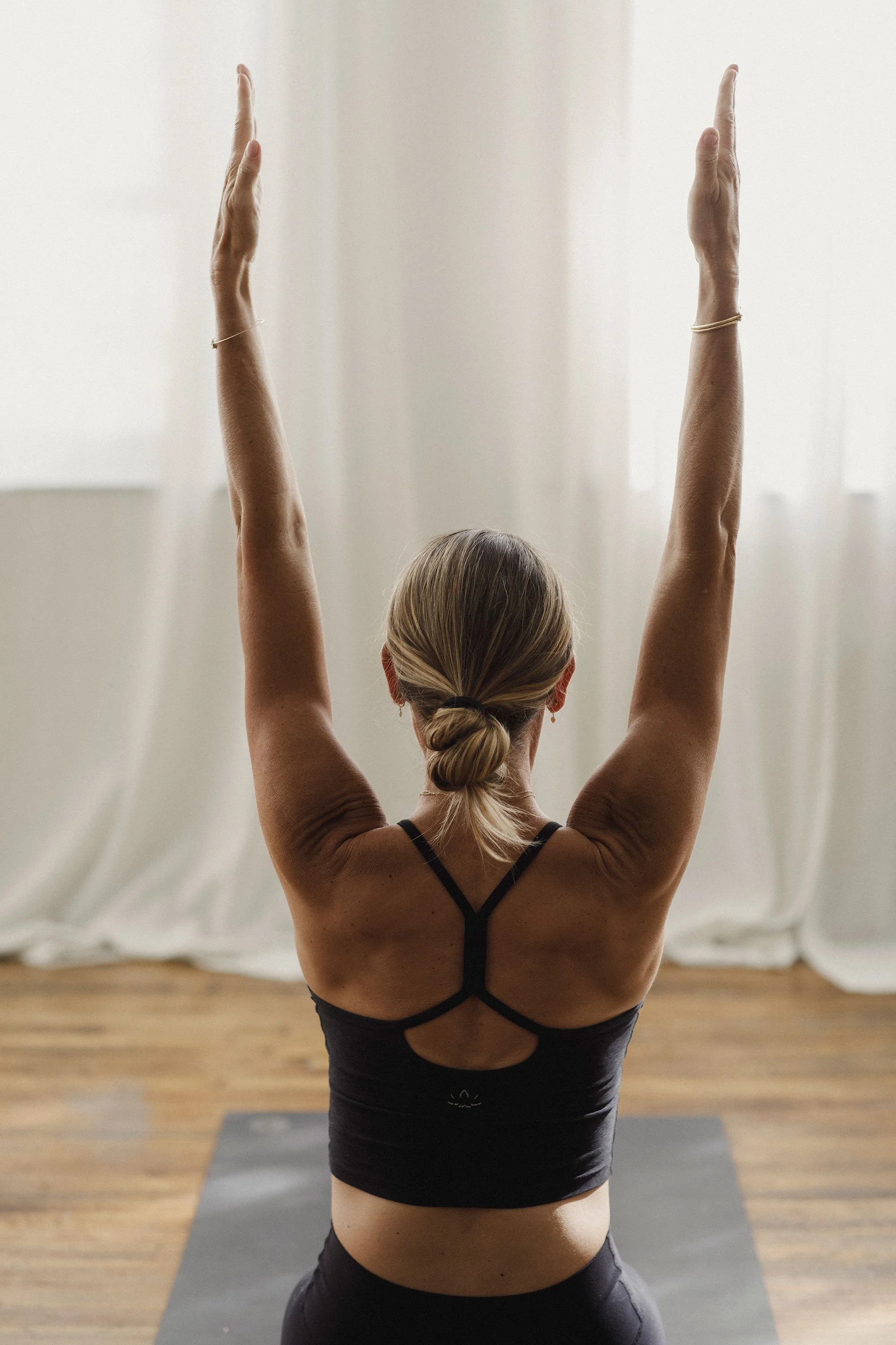 Back of woman practicing yoga, arms raised, hands together overhead, in a well-lit room with white curtains and wooden floor.